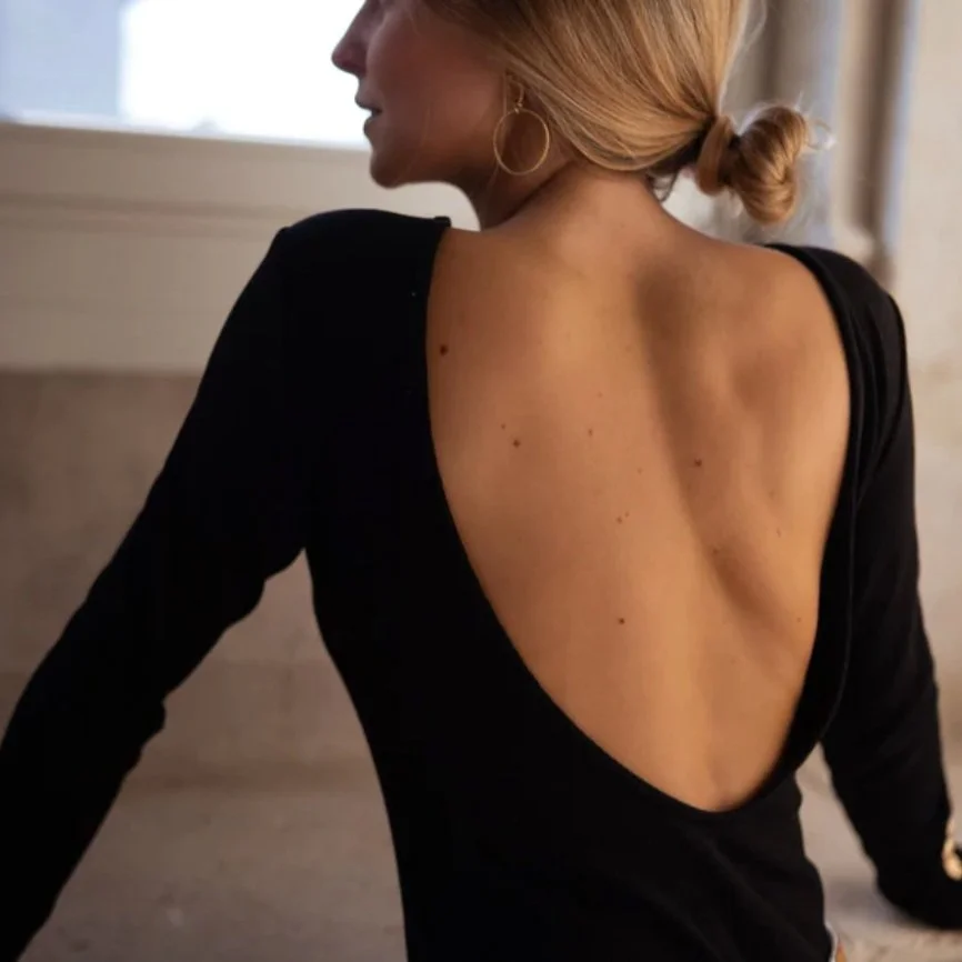 A woman with blonde hair styled in a low bun, wearing large hoop earrings and a black top with an open back, sitting indoors near a window.