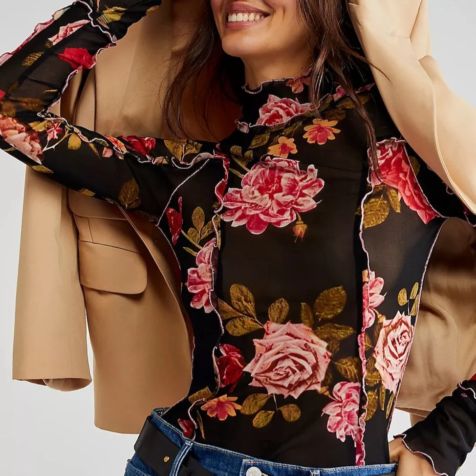 A woman wearing a floral sheer black top and a beige blazer, smiling with her hand near her head.