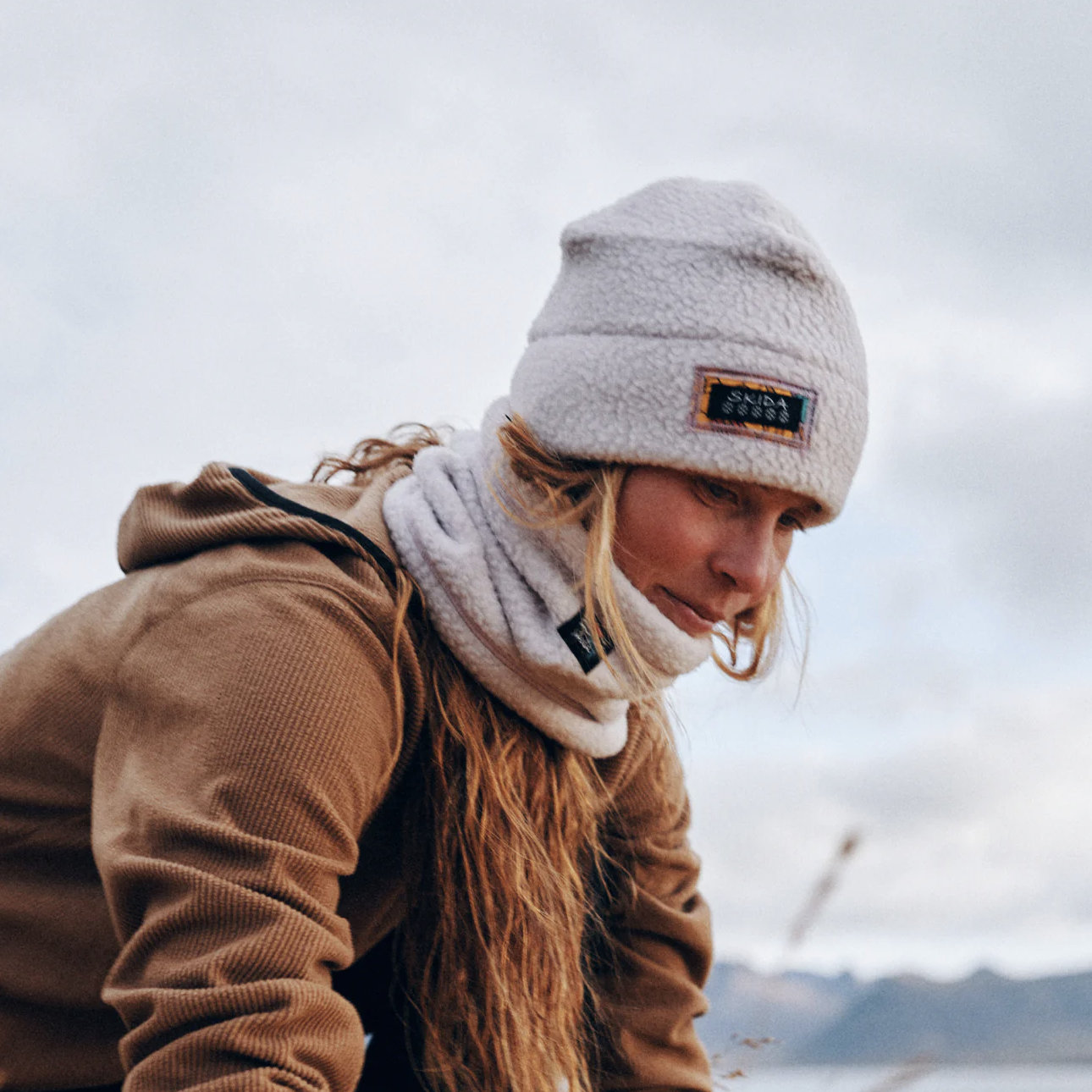 Woman wearing a white beanie and a tan jacket outdoors on a cloudy day.