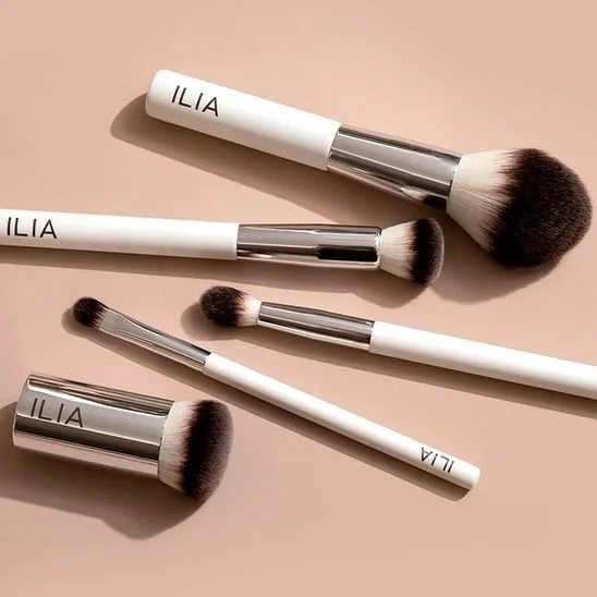 Collection of five Ilia makeup brushes on a light pink background, with two of the brushes in white and silver handles, and three smaller brushes with white handles and silver ferrules.