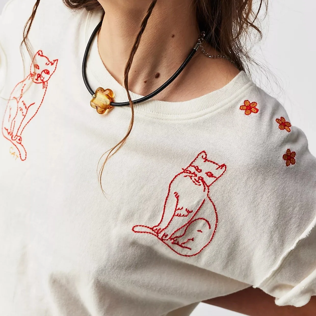 Close-up of a person wearing a white shirt with red embroidered outline of a sitting cartoonish cat and small red flowers; person also has a black choker with a gold flower-shaped pendant.