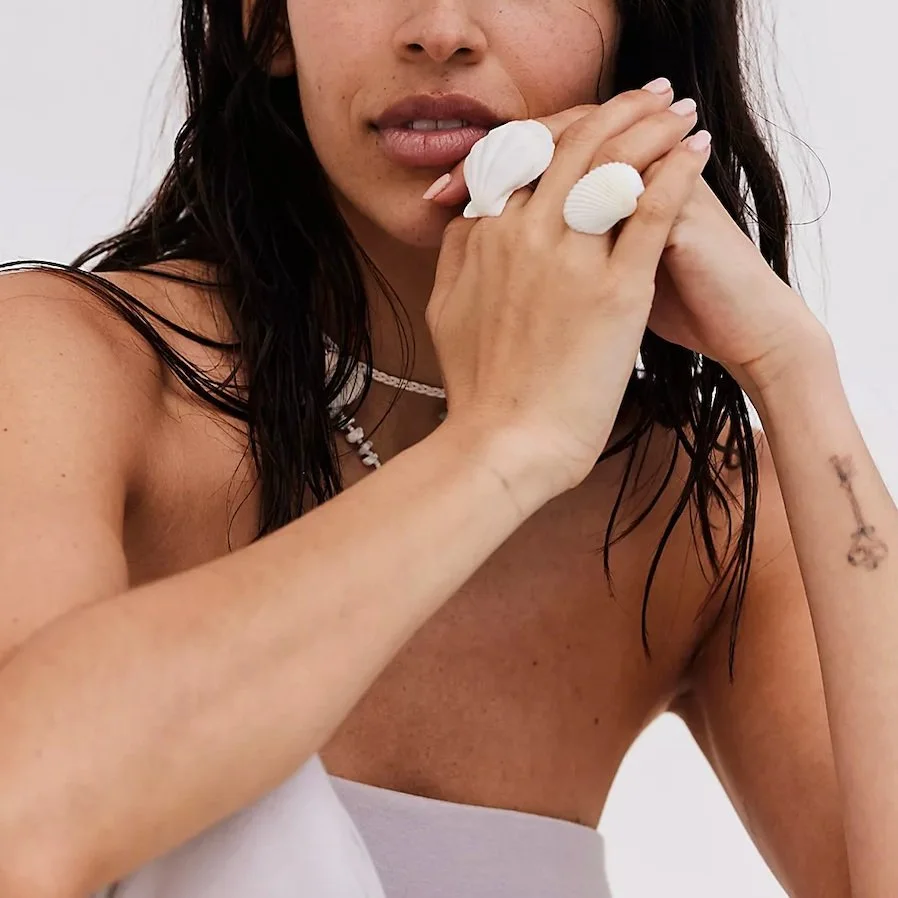 Close-up of a woman with wet dark hair, holding seashells to her lips, wearing a white strapless top and jewelry, with a tattoo on her left forearm.
