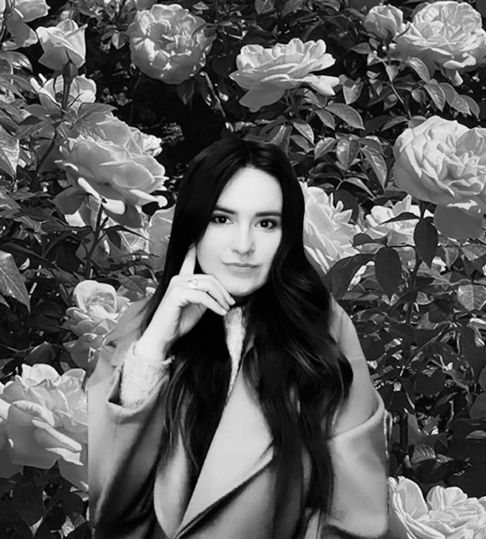 A black and white photo of a woman with long dark hair standing in front of a bush with large roses. She is resting her chin on her hand, smiling softly, and wearing a coat with her other hand inside the coat pocket.