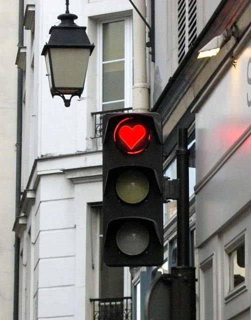 A traffic light with a red heart symbol in place of the usual red light, positioned on a city street with buildings and a street lamp in the background.