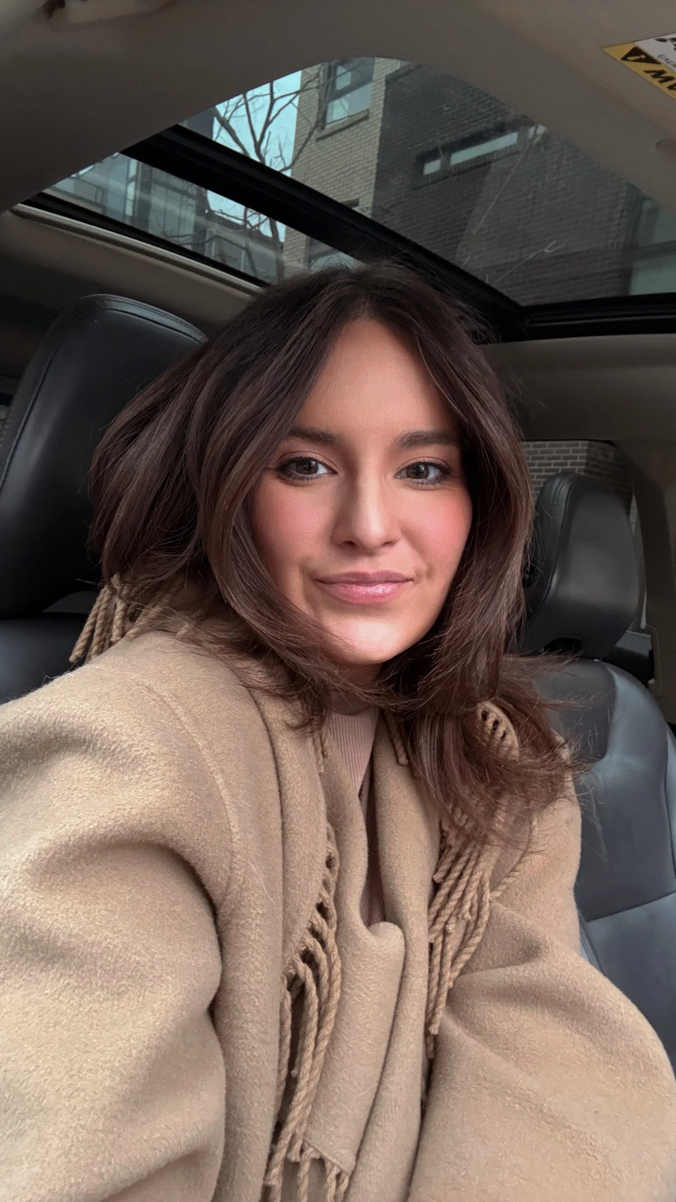 A young woman with shoulder-length brown hair and light makeup, smiling softly, sitting inside a car with a leather seat and a sunroof, with buildings visible through the window.