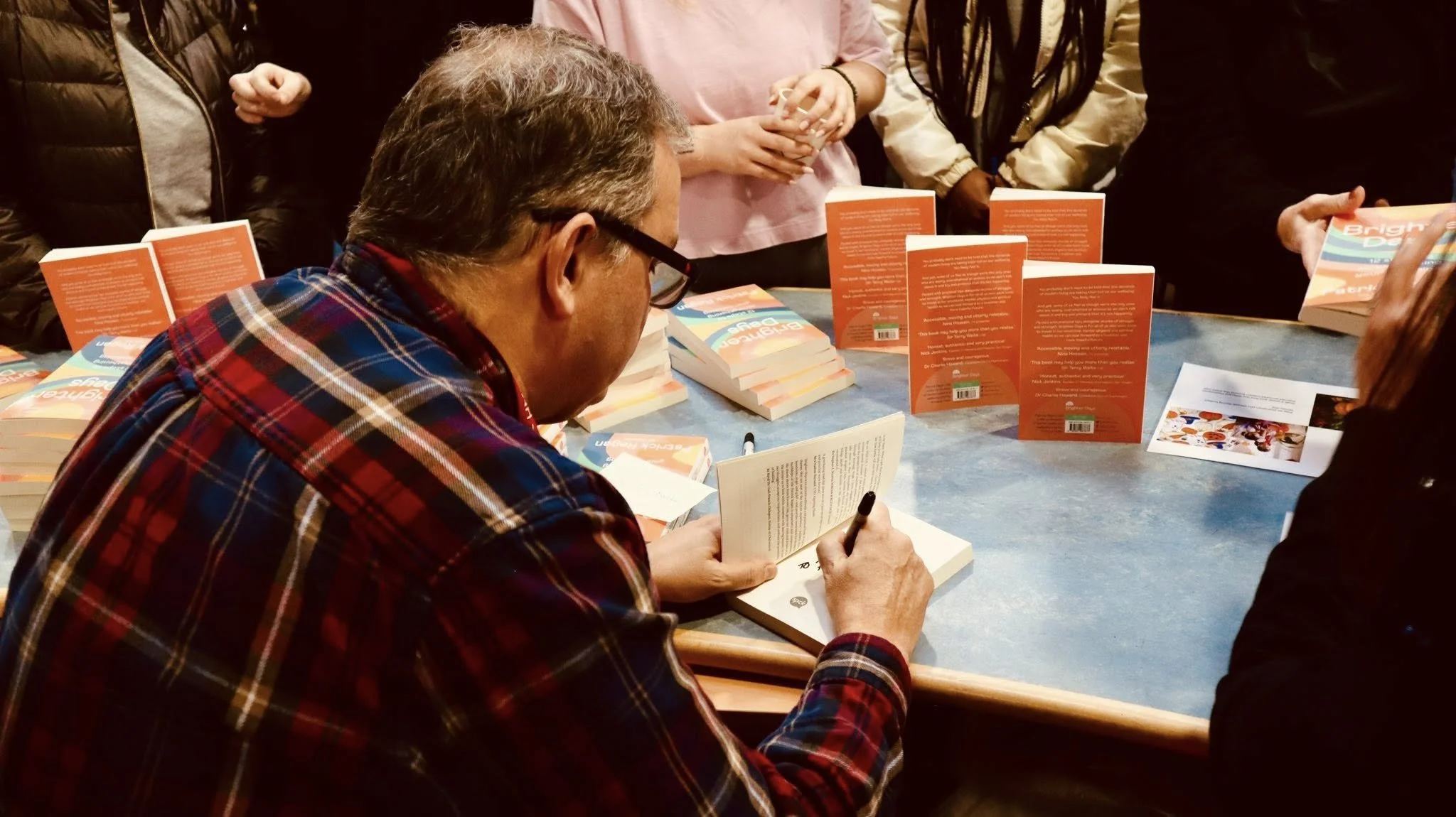 A man with glasses and a plaid shirt signing a book at a table surrounded by copies of his book and people waiting in line.
