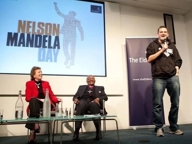 People on stage celebrating Nelson Mandela Day, including a woman in a red blazer, a man in a black blazer, and a young man with a microphone. A large screen behind displays a Nelson Mandela silhouette and text reading "Nelson Mandela Day."