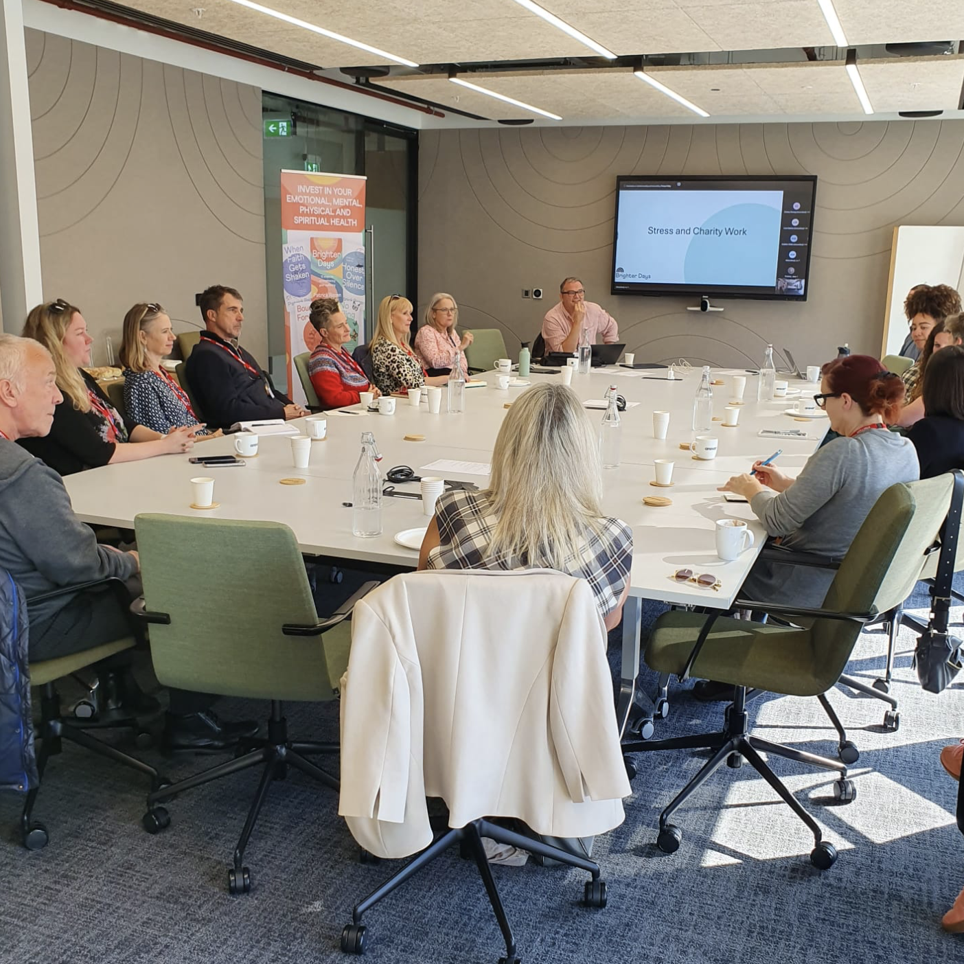 People attending a meeting in a conference room, with a presenter and a large screen displaying a presentation on stress and charity work.