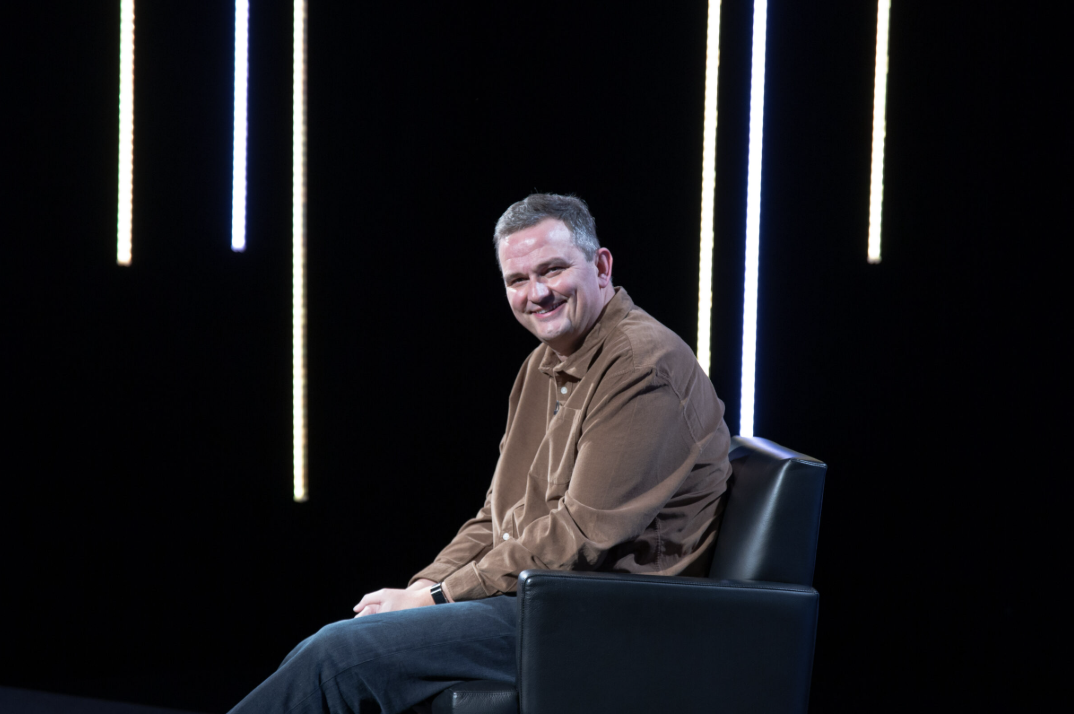 A man in a brown shirt sitting on a black chair against a black background with vertical light strips.