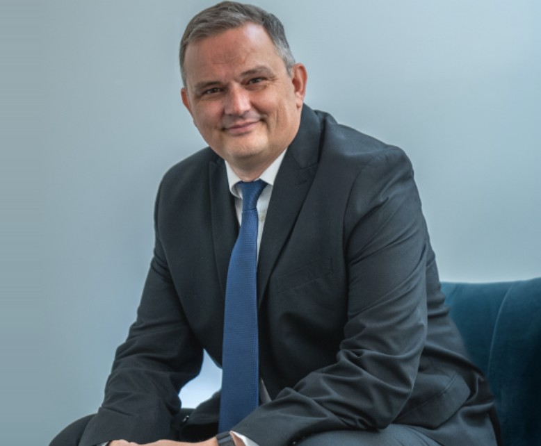 A man in a dark suit, white shirt, and blue tie sitting on a blue chair against a light blue background, smiling at the camera.