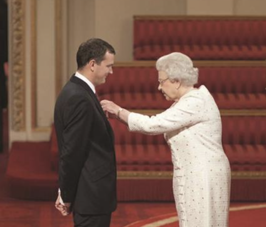 Queen Elizabeth II presenting an award or honor to a young man in a formal setting with red carpeted stairs in the background.