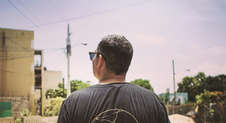 Man with dark hair and sunglasses facing away outdoors on a sunny day, with buildings, trees, and power lines in the background.