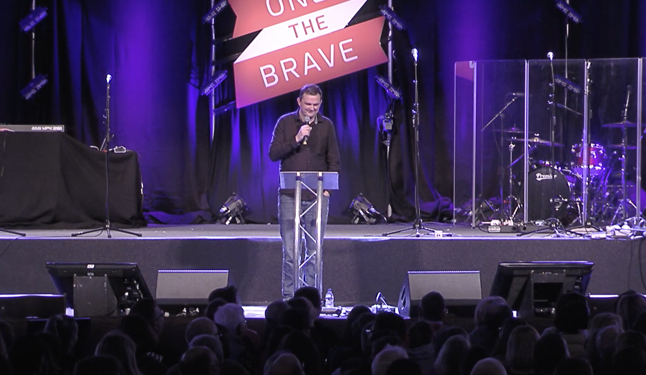 Man speaking on stage at a conference or event with a sign that reads 'One of the Brave' behind him, stage lighting, microphones, musical instruments, and audience in the foreground.