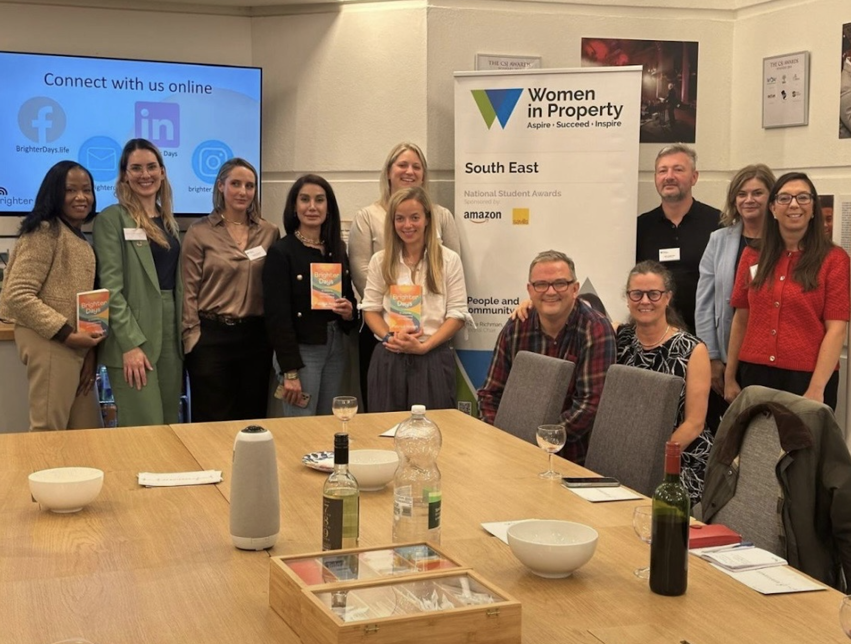 A group of people posing for a photo at a gathering or event, with a banner and a screen in the background. Several individuals are holding copies of a book titled 'Brighter Days.' The table in the foreground has bottles, bowls, and glasses.