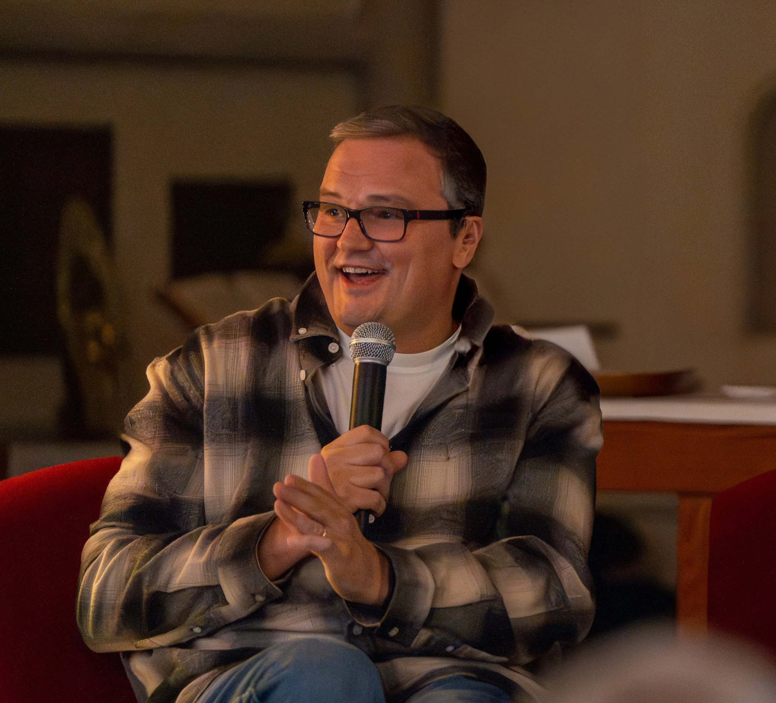 A man wearing glasses and a plaid shirt holding a microphone, smiling while speaking, in a cozy indoor setting.
