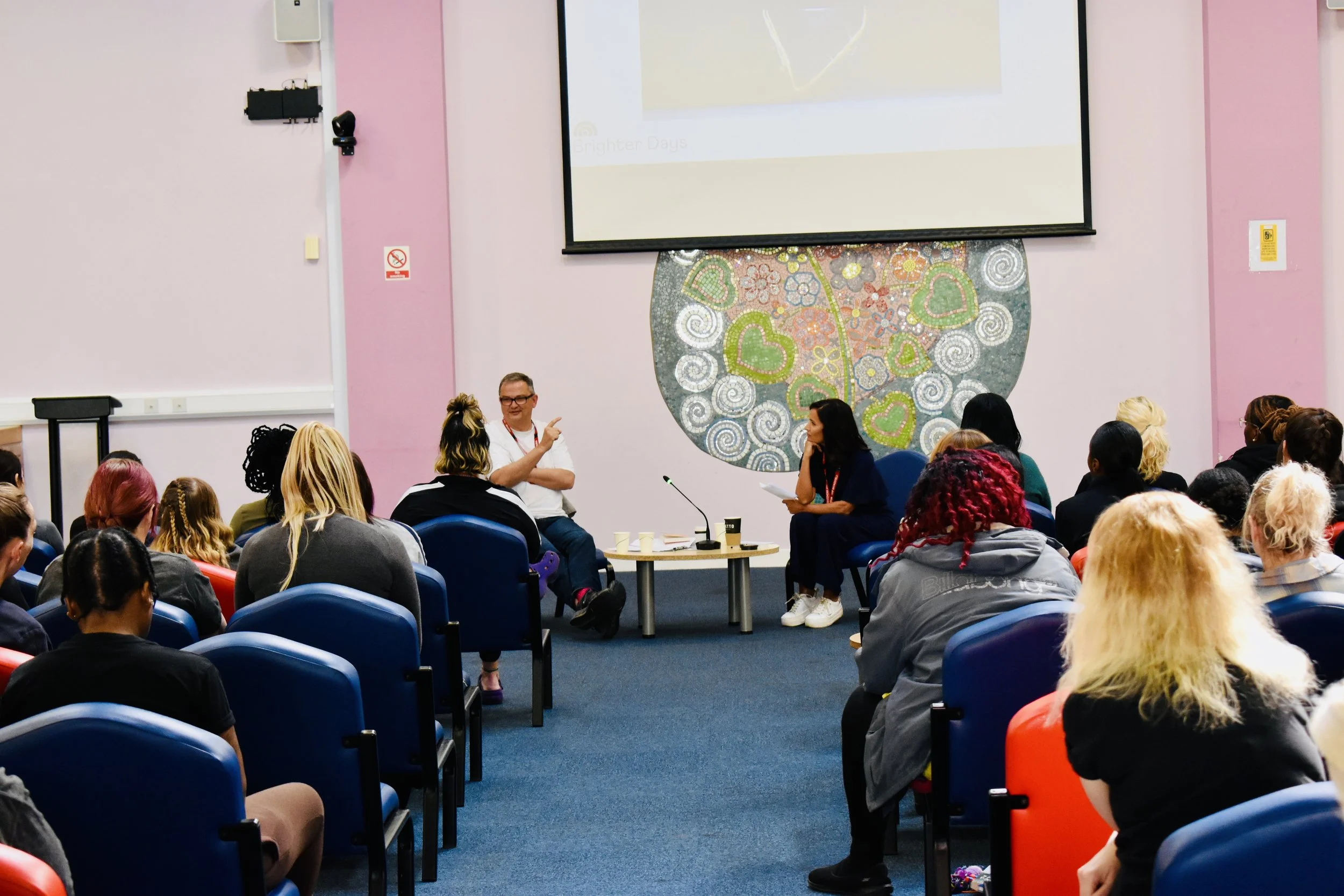 A group of people attending a seminar or panel discussion in a conference room with pink walls and a large colorful wall art piece behind the speakers. One man is speaking while a woman listens and takes notes.