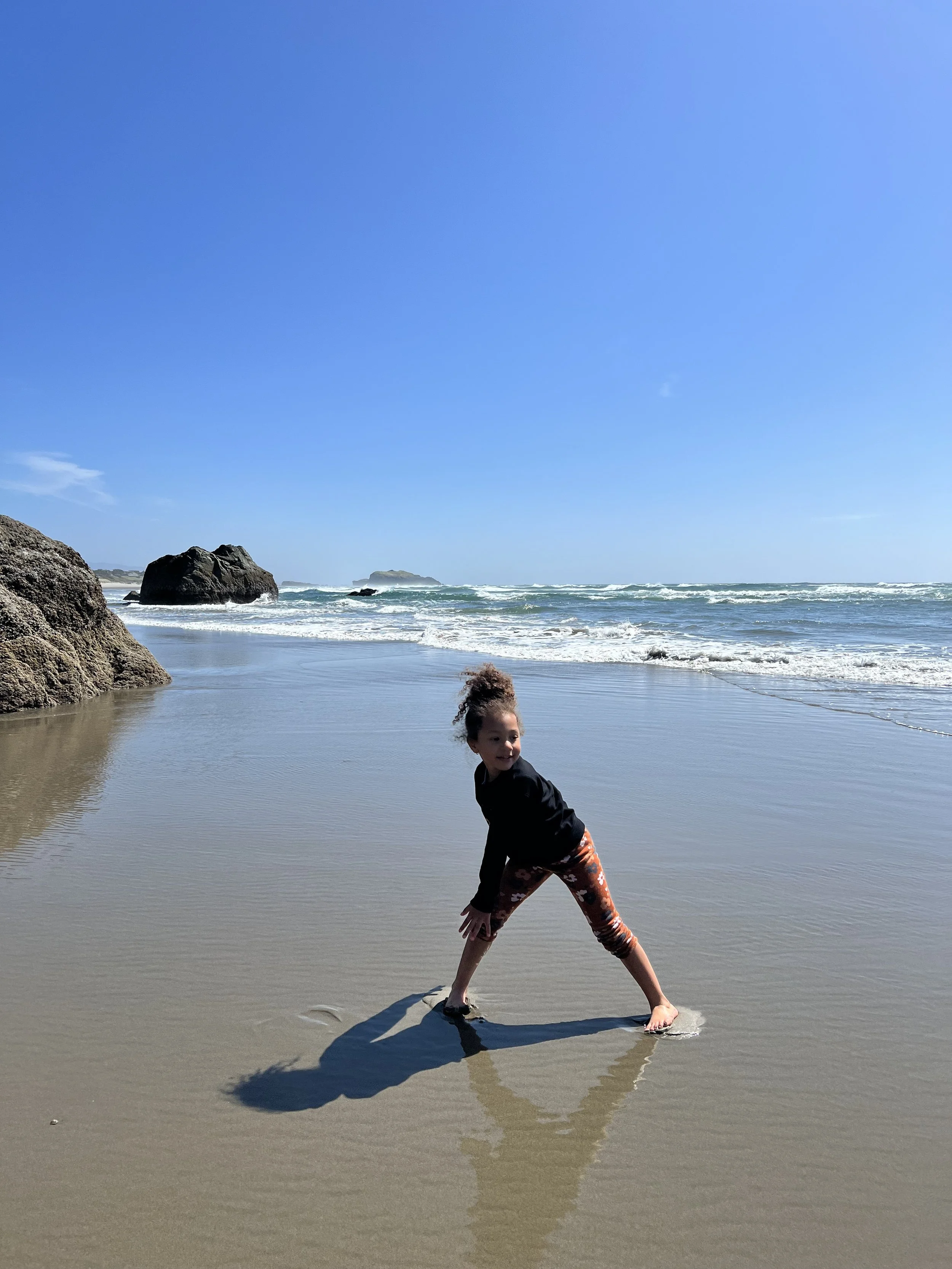 A young girl playing on a sandy beach near the ocean with rocks in the background, under a clear blue sky.