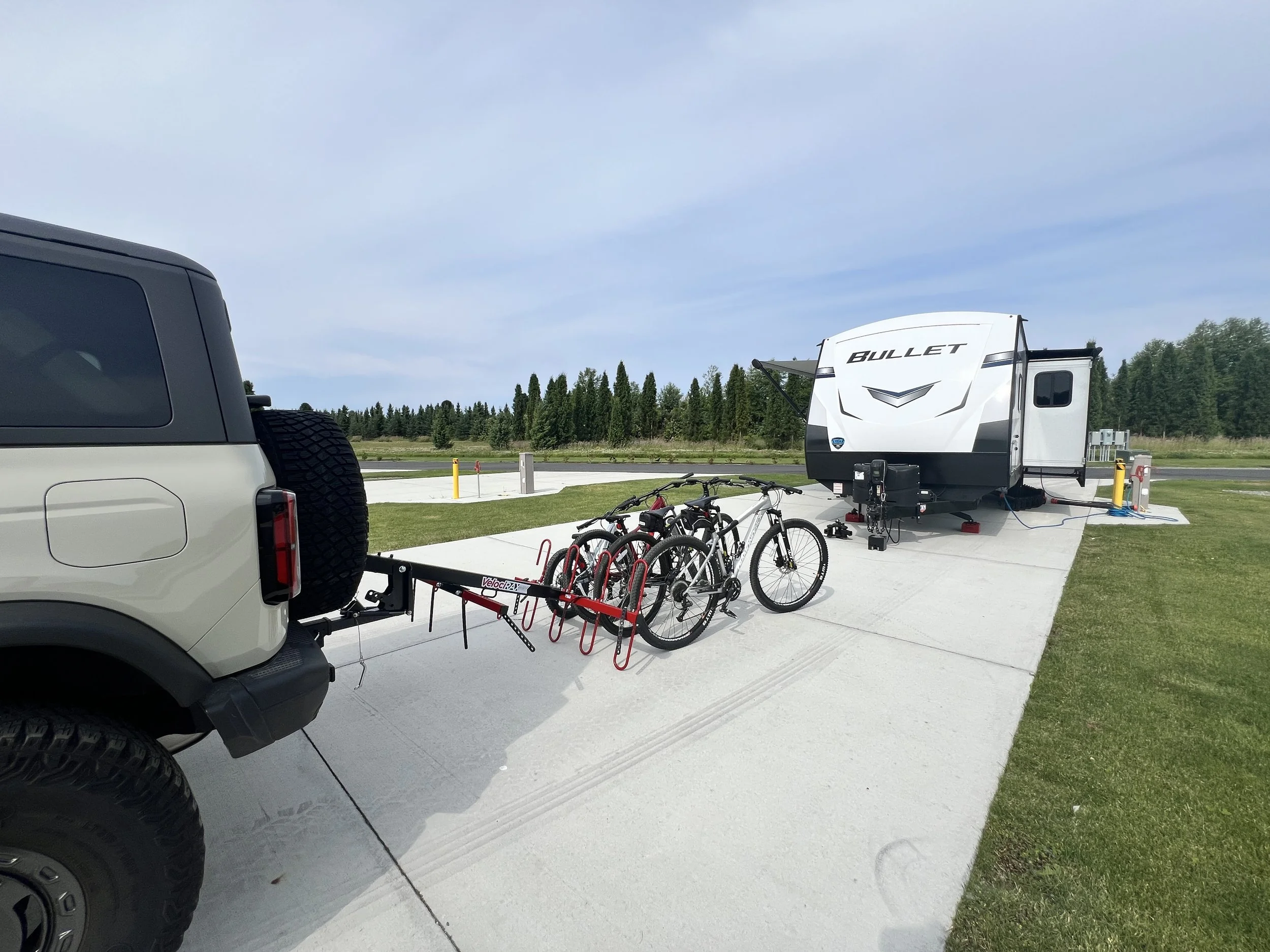 A white and gray SUV towing a bike rack with four bicycles parked in front of a travel trailer with the word "BULLET" on it, at a campsite with a concrete pad, green grass, and trees in the background.
