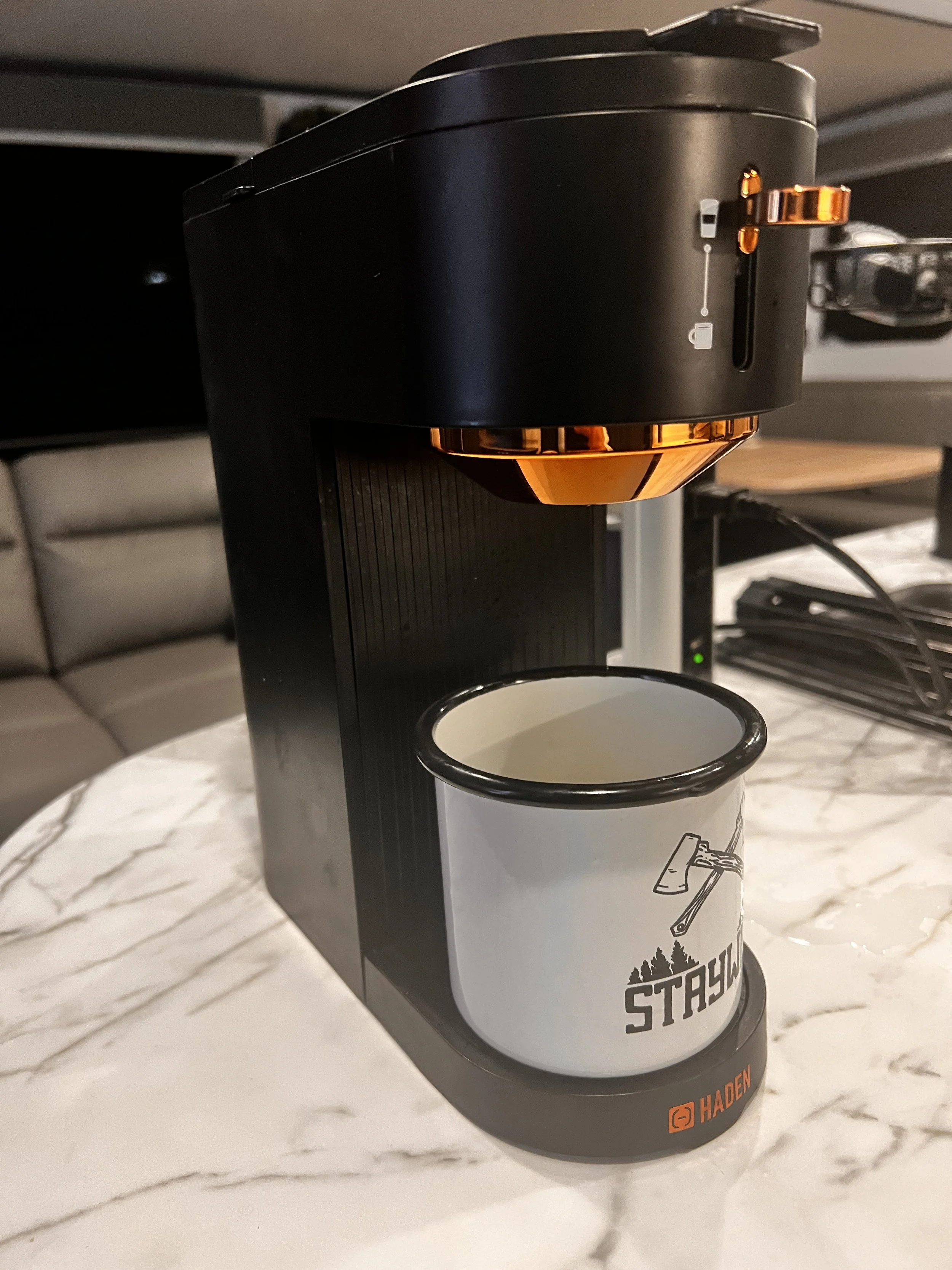 A coffee maker brewing into a white mug with black rim and a STAYWILD logo, placed on a white marble countertop.