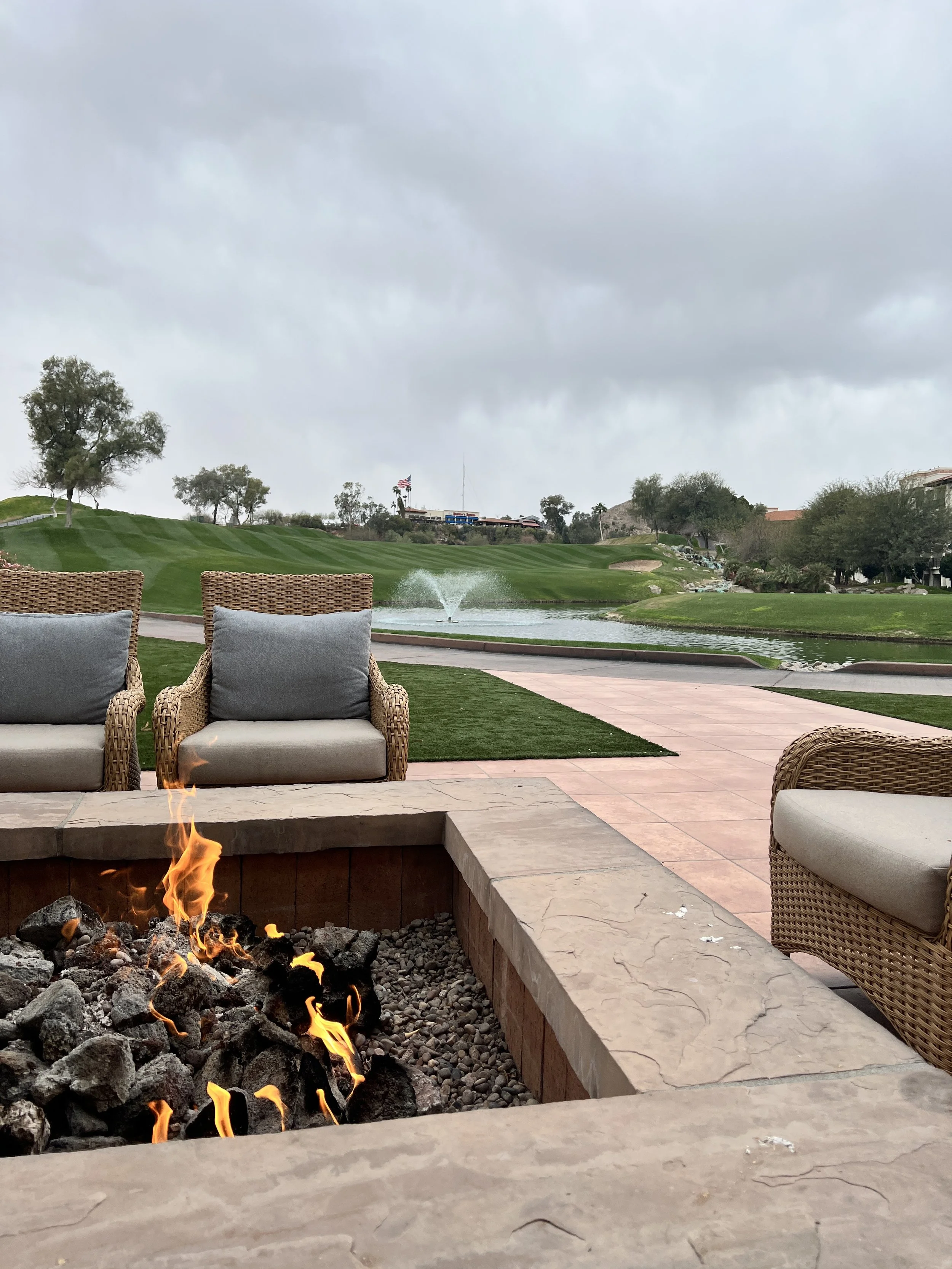 Outdoor seating area with wicker chairs and gray cushions, a fire pit with flames, and a golf course with a pond and fountain in the background under cloudy sky.