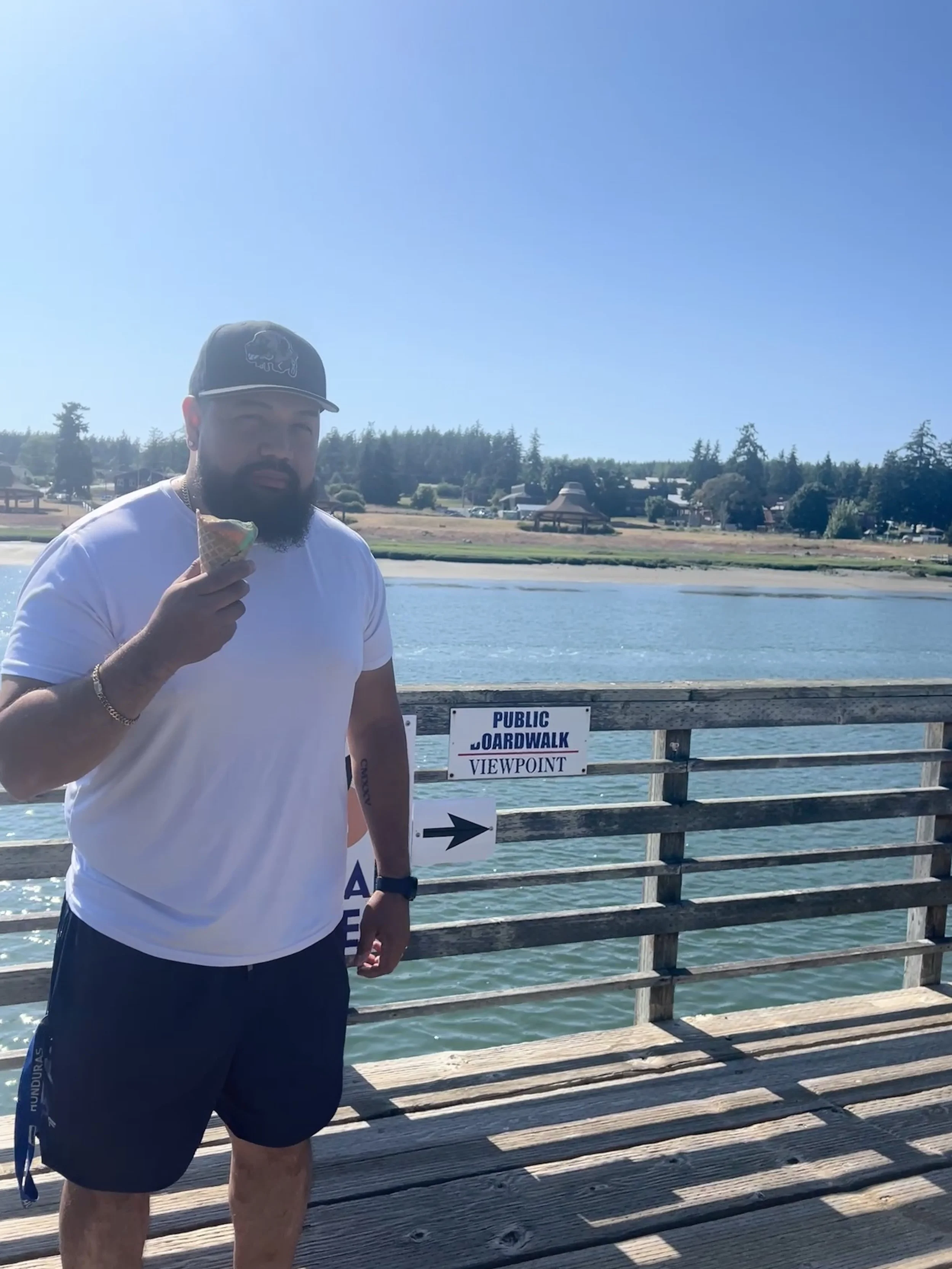 A bearded man in a white t-shirt and black shorts holding an ice cream cone standing on a wooden dock by the water, with a sign that reads 'Public Boardwalk Viewpoint' and an arrow pointing to the right.