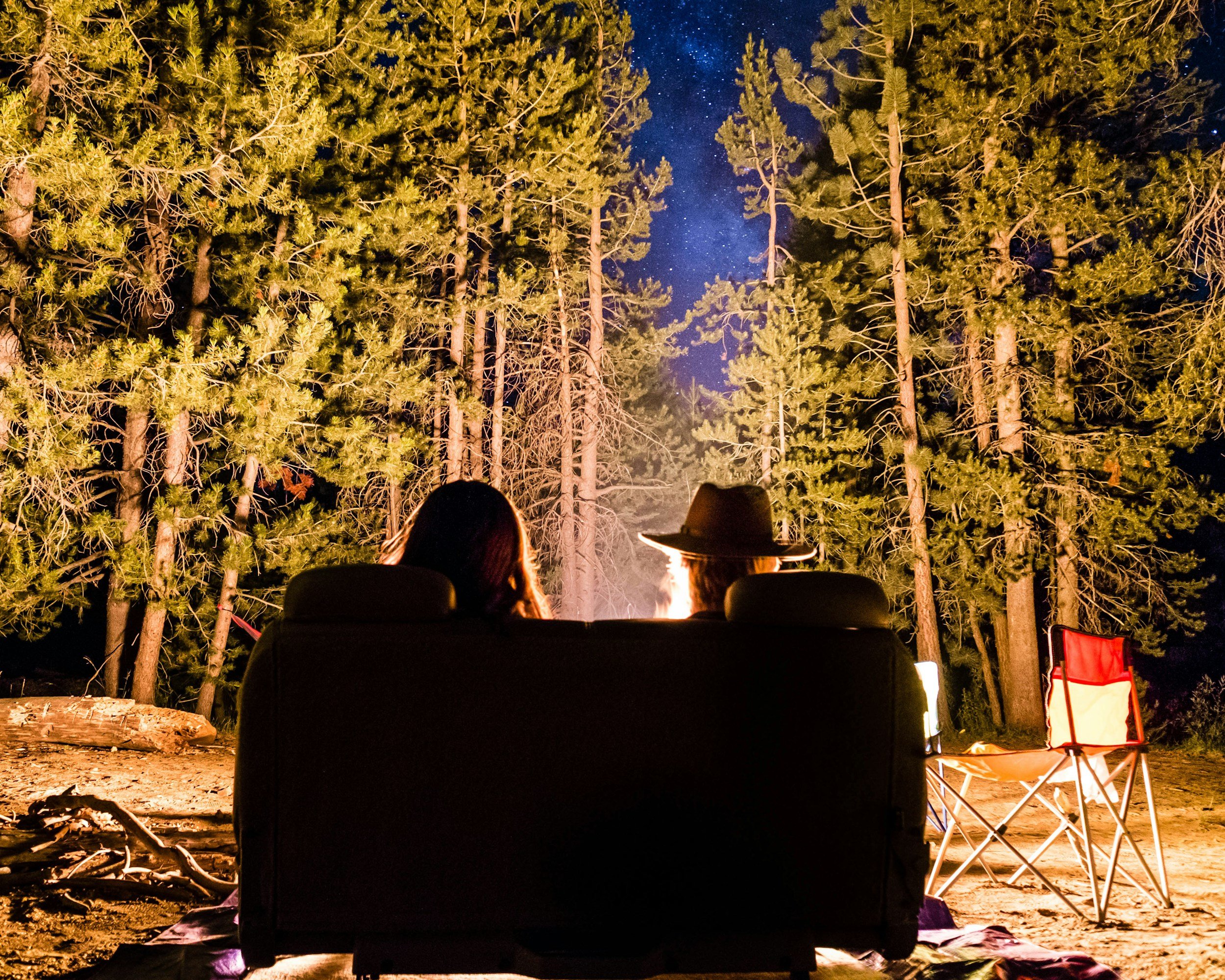 Two people are sitting on a couch around a campfire in a forest at night. One person is wearing a hat, and a red and white camping chair is placed nearby. The scene is illuminated by the campfire, with tall trees and a starry sky in the background.