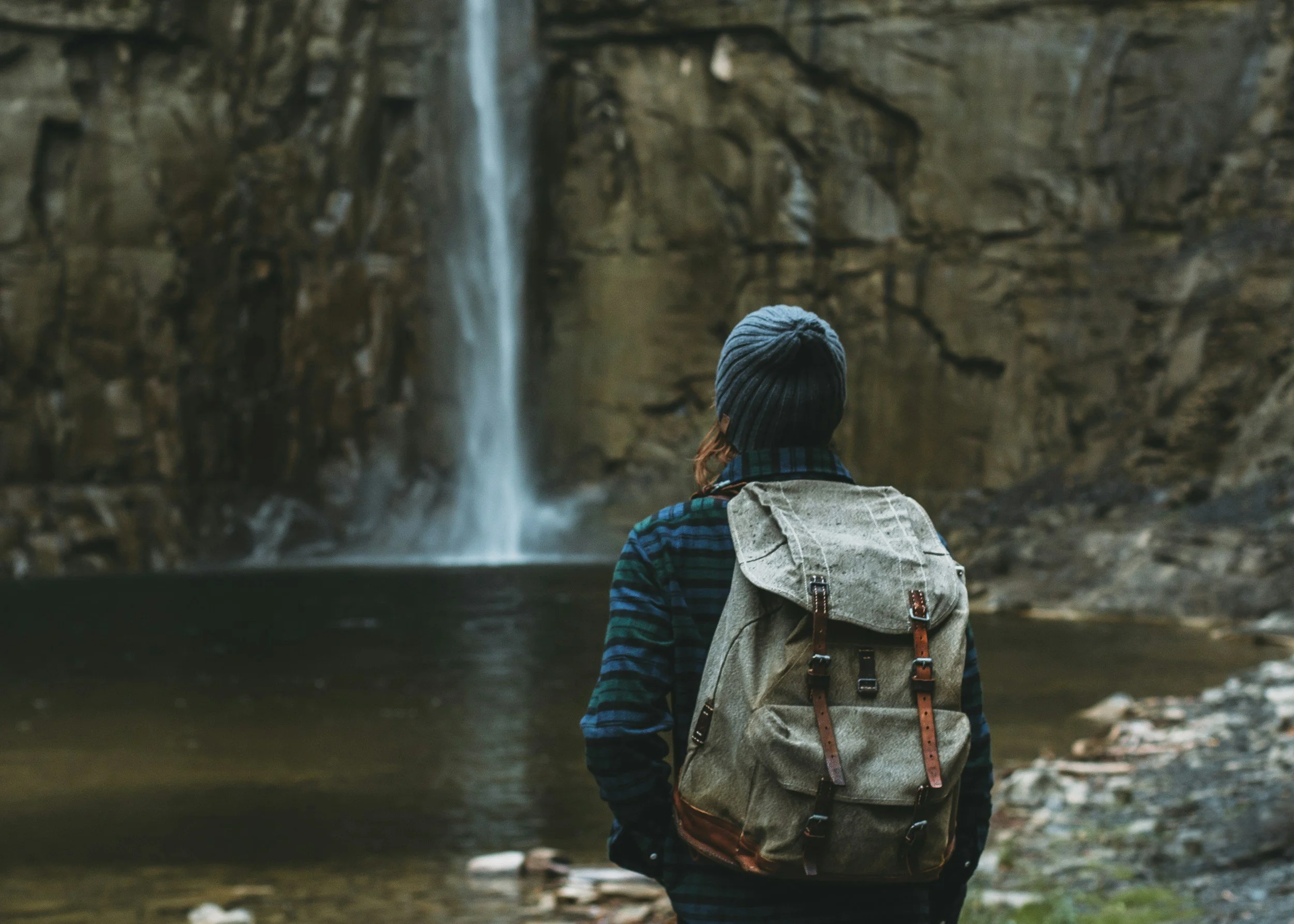 Person with a backpack standing near a waterfall in a rocky outdoor setting