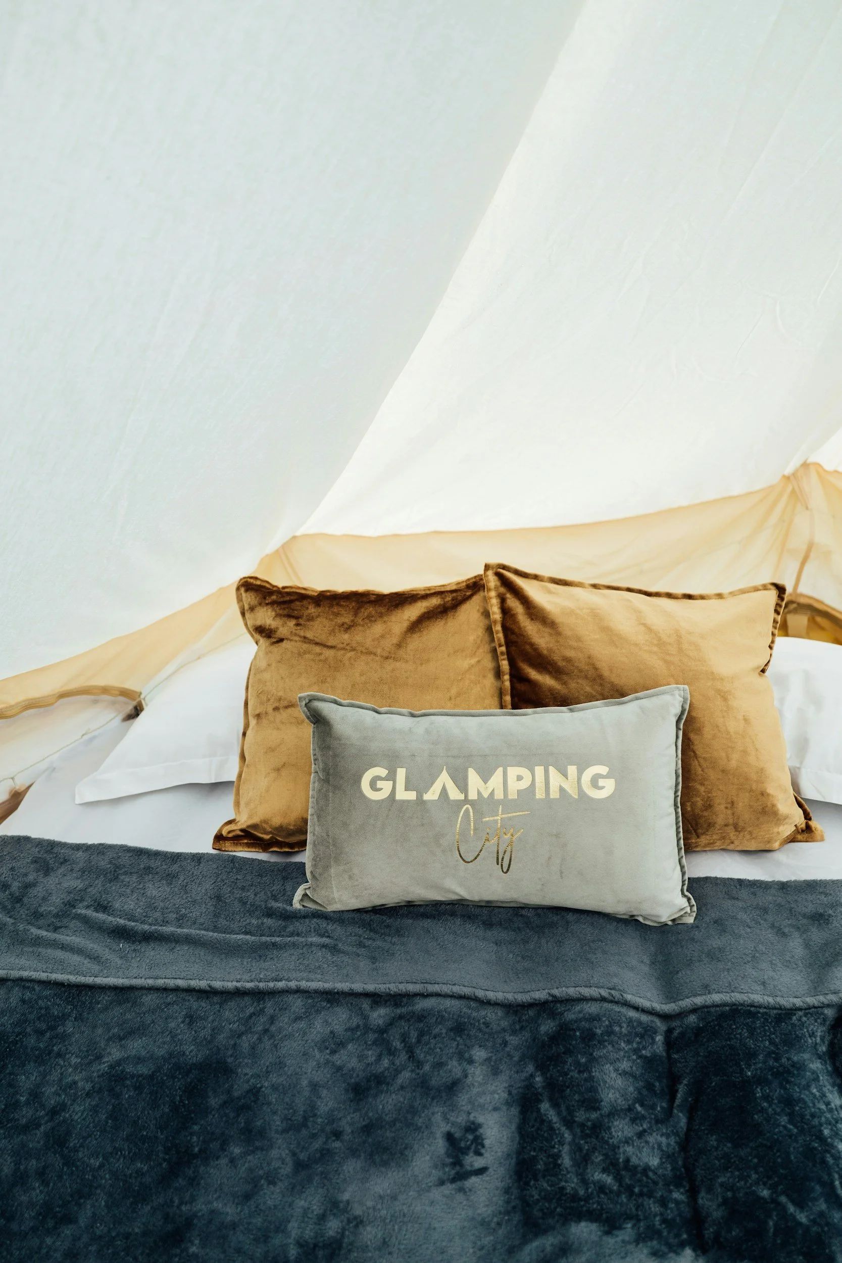 Bed with brown and silver pillows, one pillow reading 'Glamping City,' in a tent with beige walls and white ceiling.