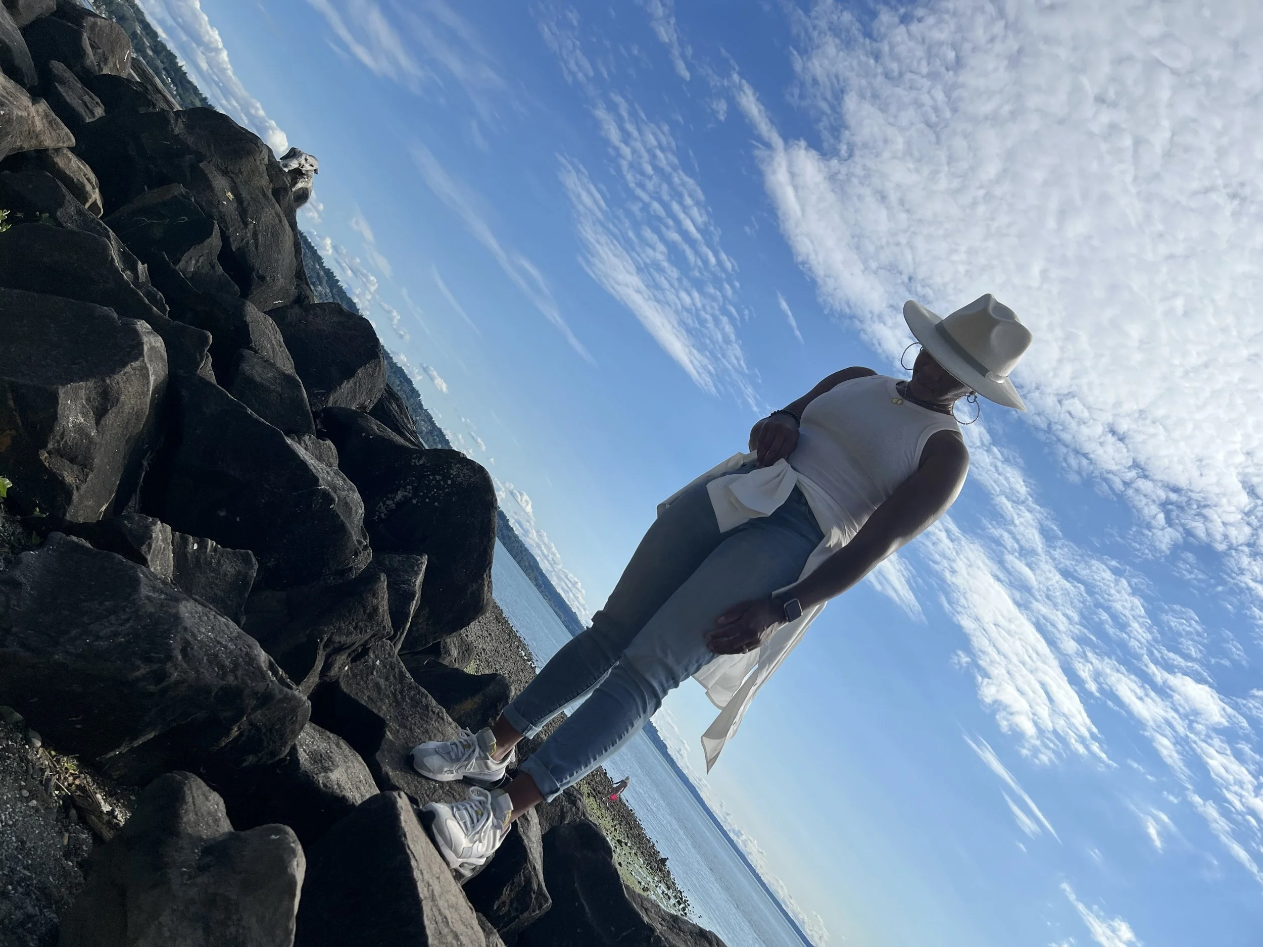A woman standing on rocks by the water, wearing a white hat, white sleeveless top, jeans, and sneakers, with a scenic sky and water in the background.