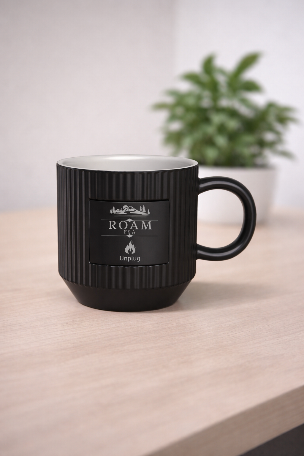 Black coffee mug with ridged texture and a label that reads "ROAM F&A Unplug" with a mountain and flame graphic, placed on a light wooden surface with a blurry green plant in the background.