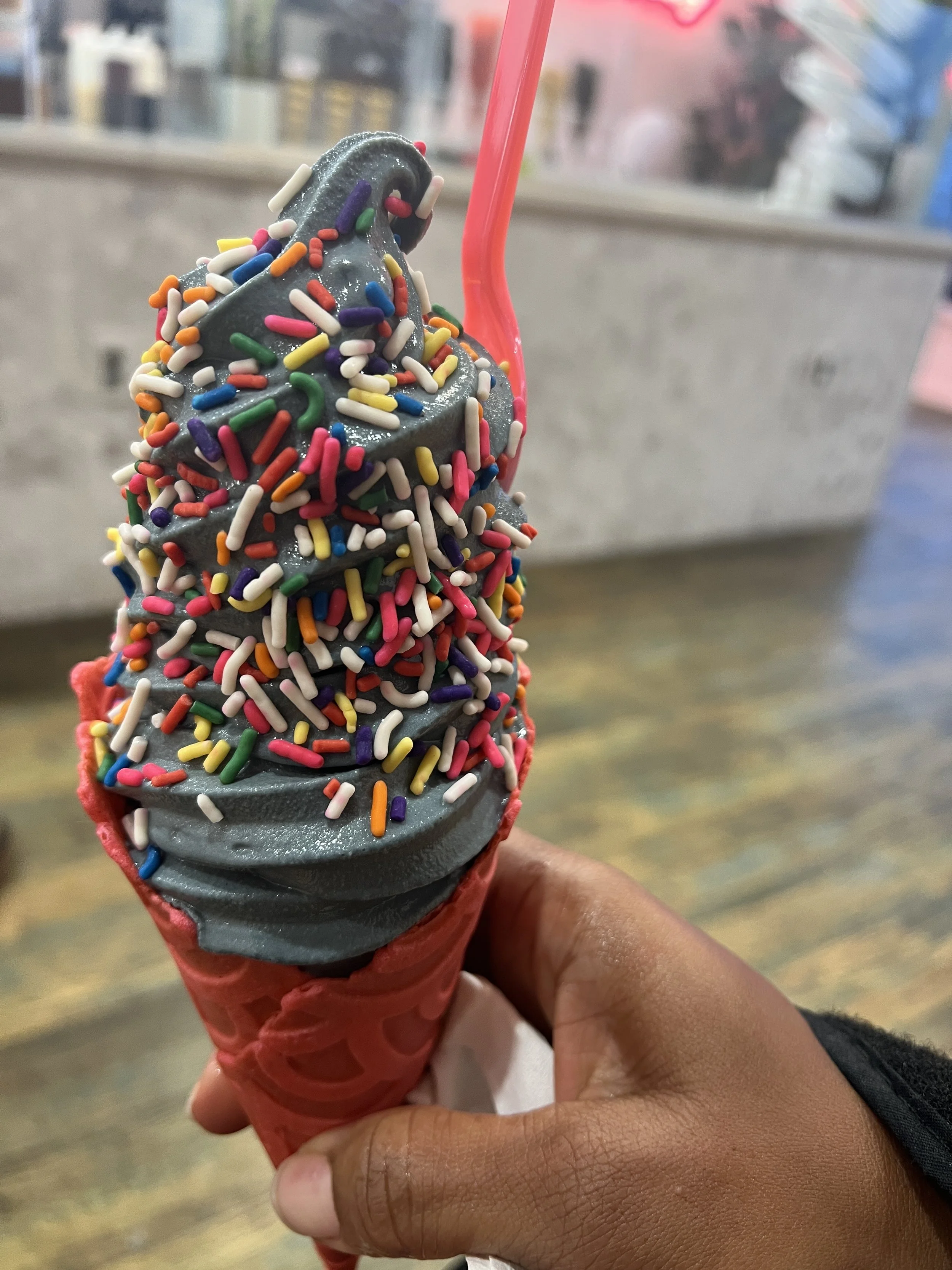 A hand holding a red cone with dark gray soft serve ice cream topped with colorful rainbow sprinkles and a pink straw.