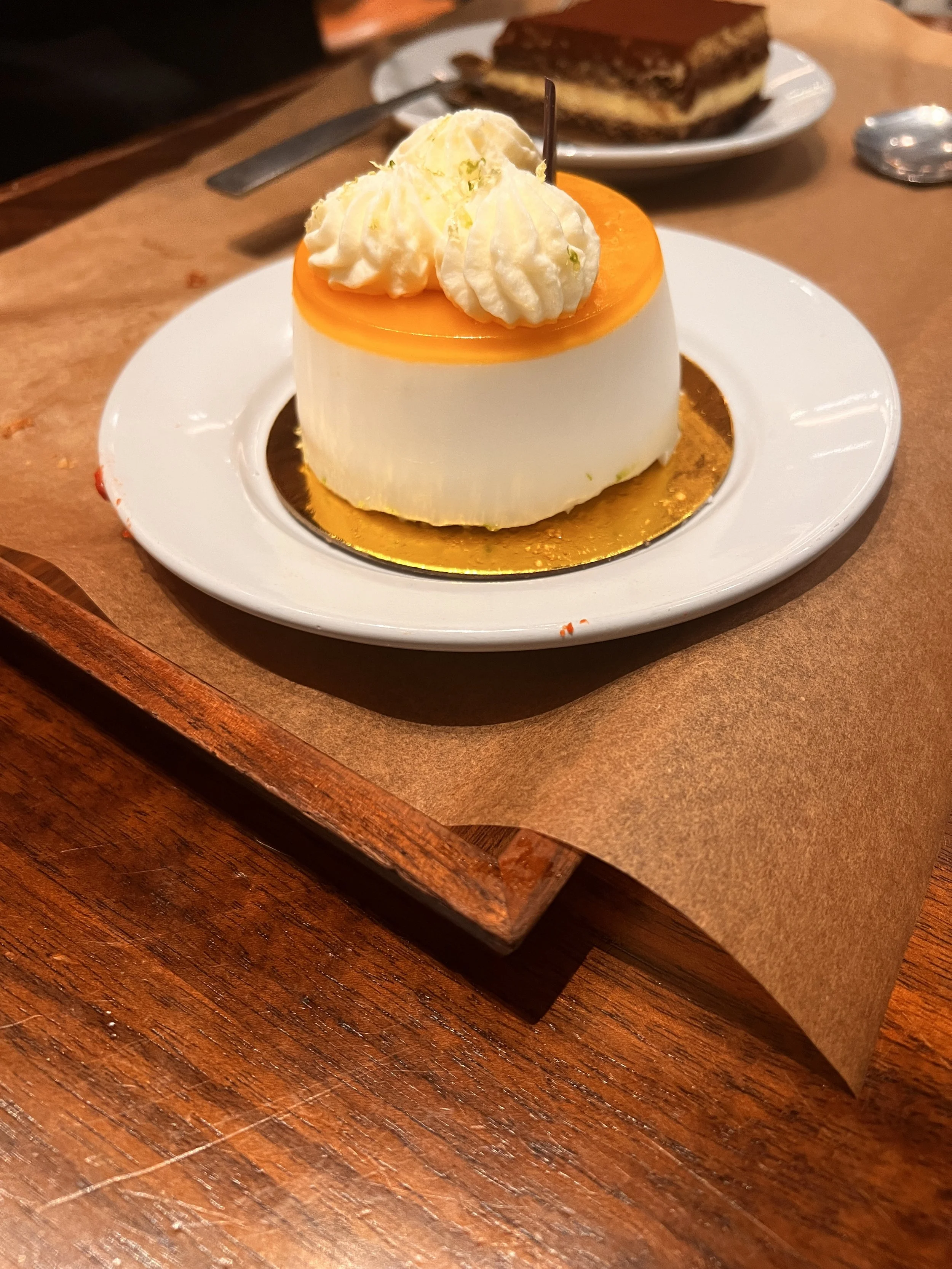 A creamy dessert topped with whipped cream and caramel sauce, served on a white plate with a slice of chocolate cake in the background.
