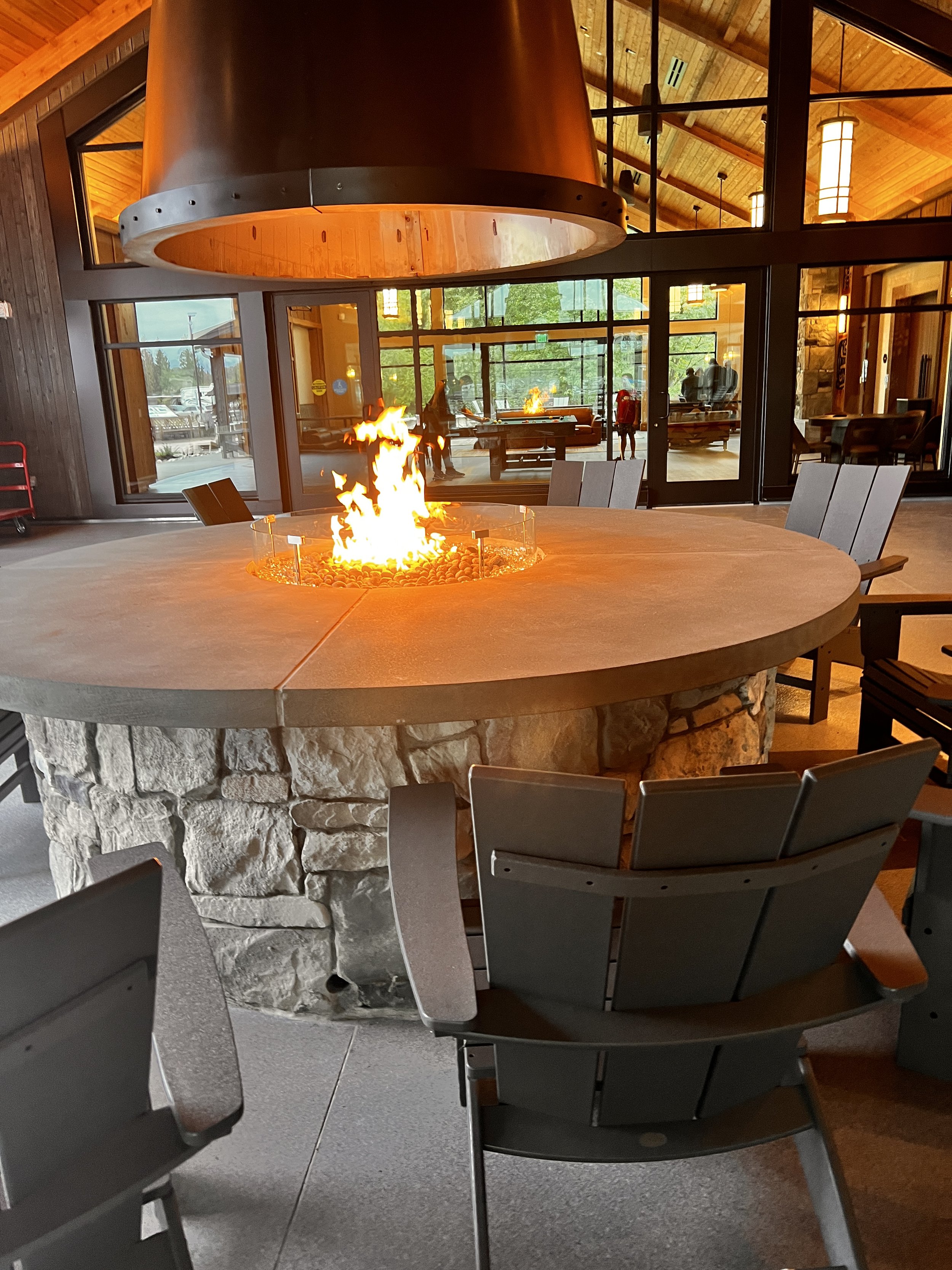 Indoor dining area with a round stone fire pit and a large metal hood above it. Chairs surround the fire pit, and glass windows and doors reveal a parking lot and trees outside.