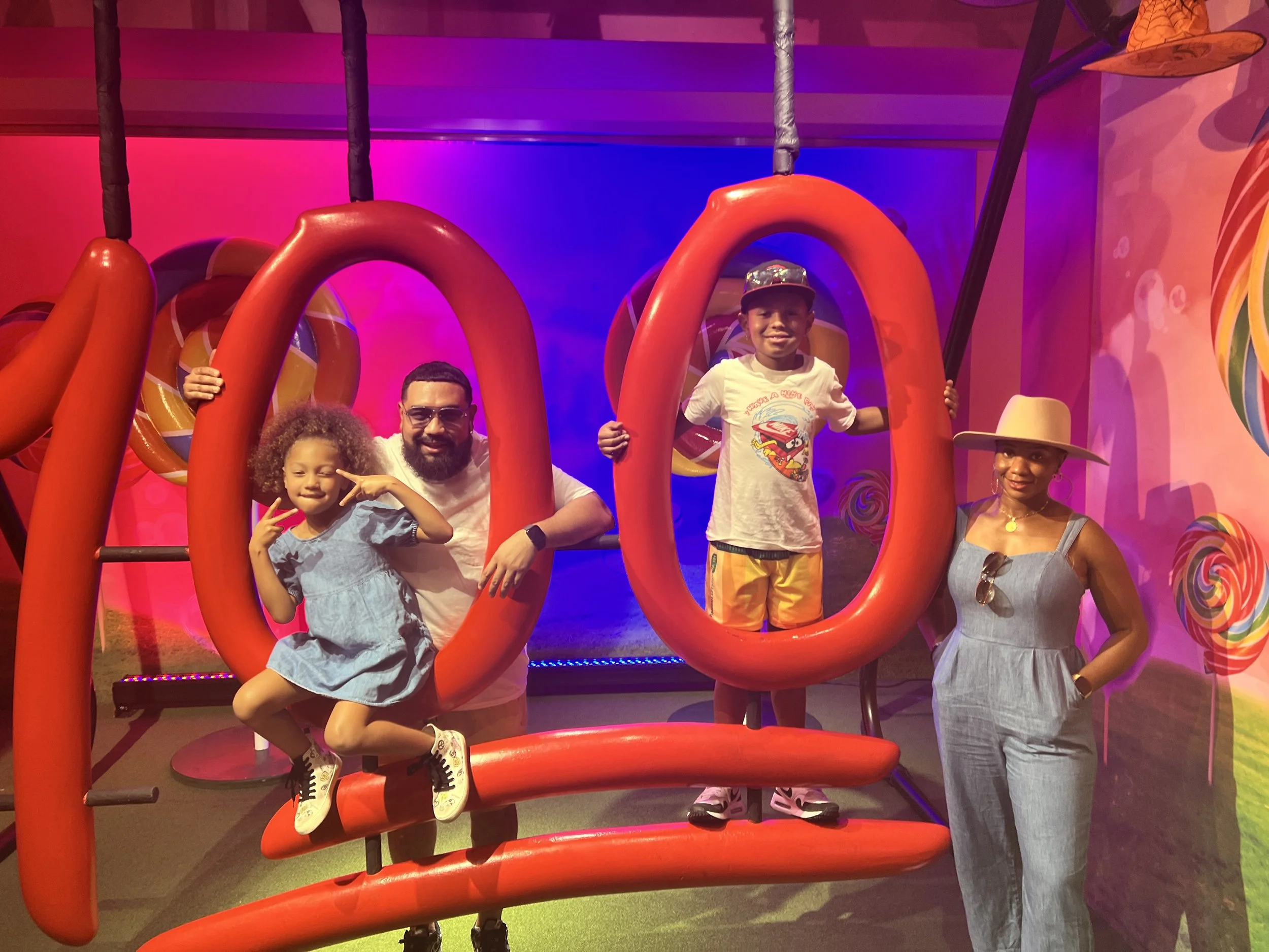 Four people enjoying a colorful indoor playground with giant red metal loops and vibrant background lighting. A young girl is sitting on one of the loops, smiling and making a peace sign. A man crouches beside her, smiling. A young boy stands inside 