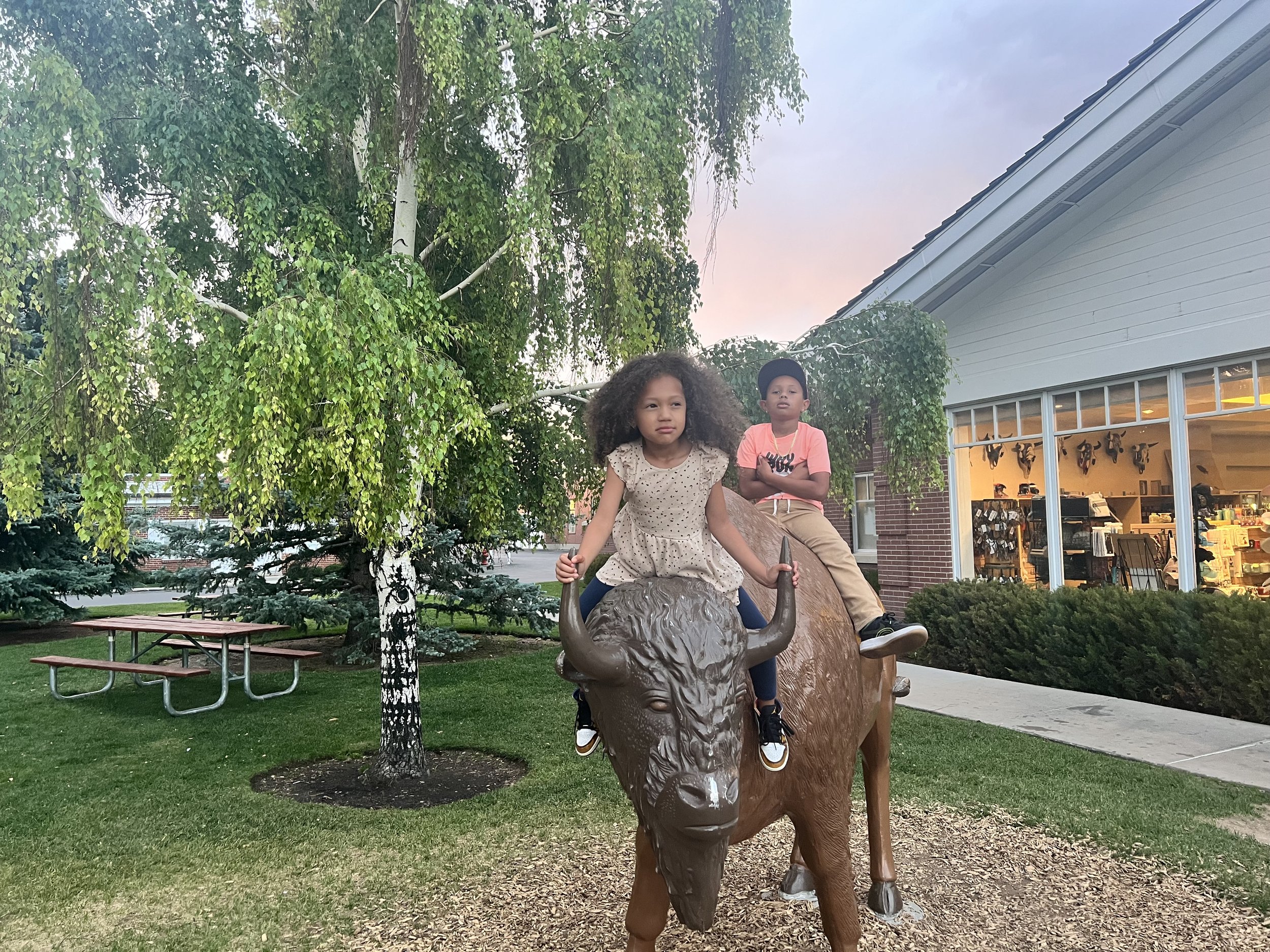 Two children, a girl and a boy, riding on a park statue of a buffalo, with trees and a building with large windows in the background.