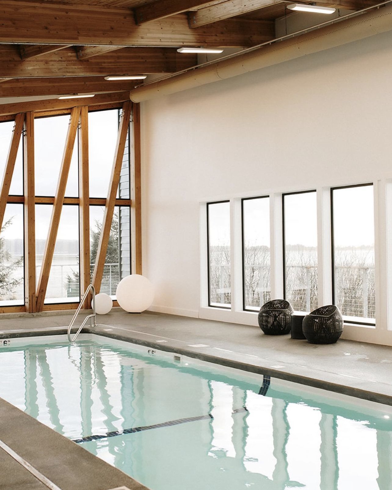 Indoor swimming pool area with large windows, wooden ceiling, and modern decor including black wicker chairs and spherical lamps.