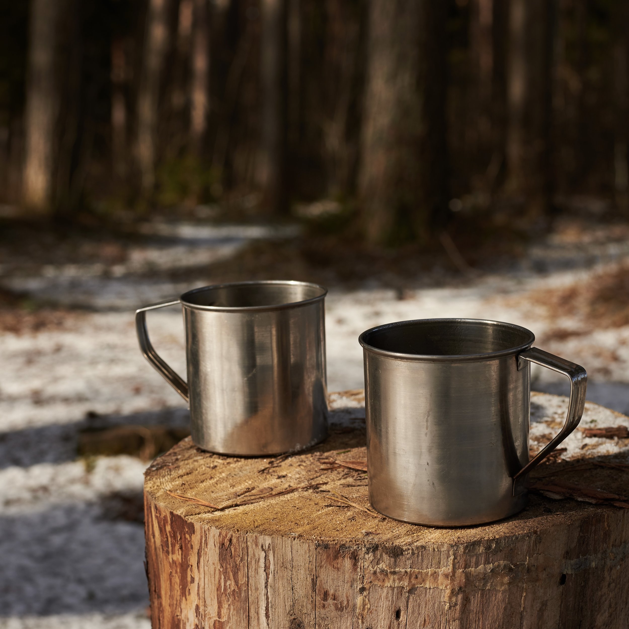 Two stainless steel mugs with handles resting on a tree stump outdoors in a forested area, with snow on the ground.