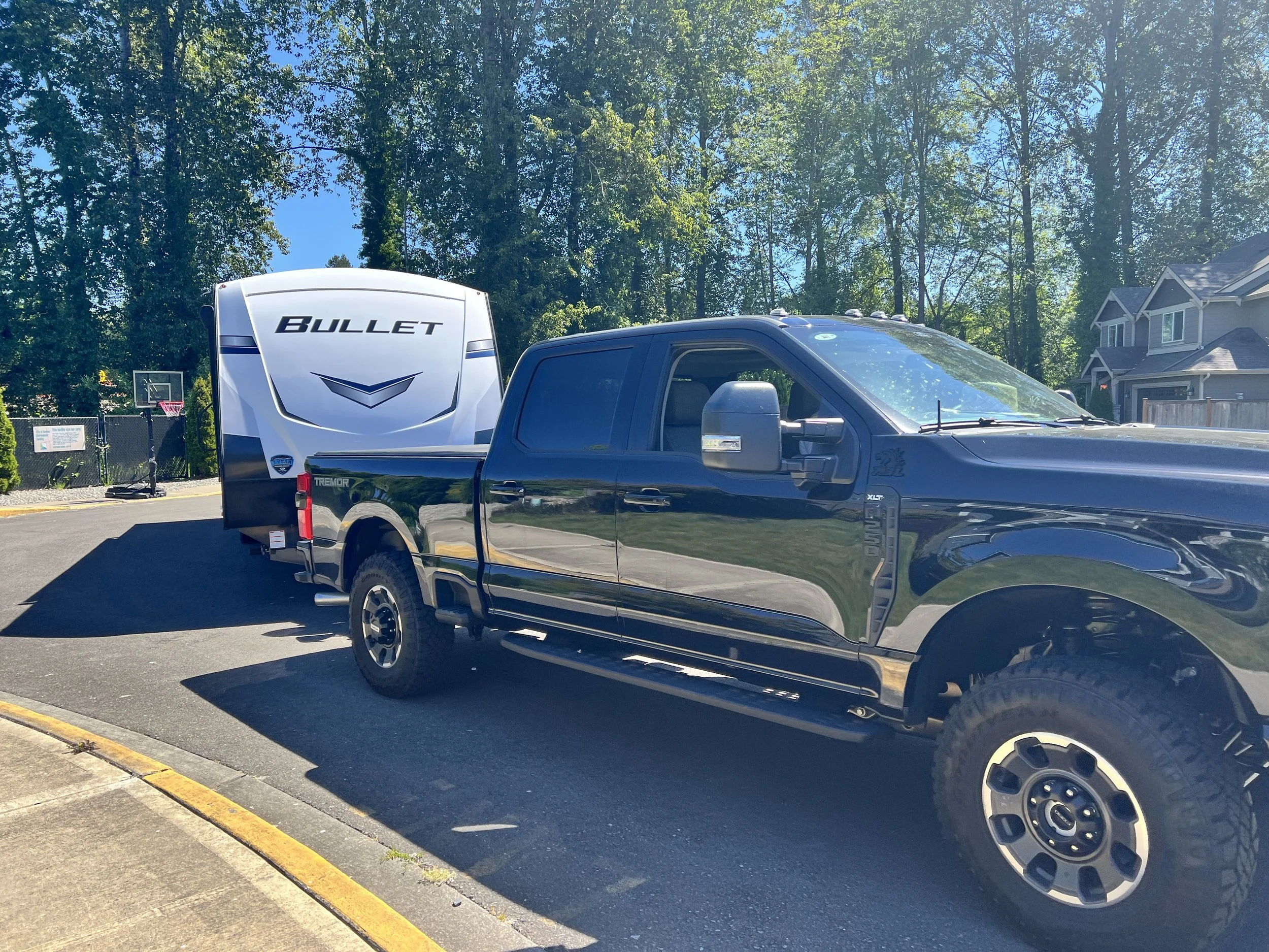A black pickup truck with large tires pulling a travel trailer with the word "Bullet" on the back, parked on a driveway in a suburban neighborhood with trees and houses nearby.