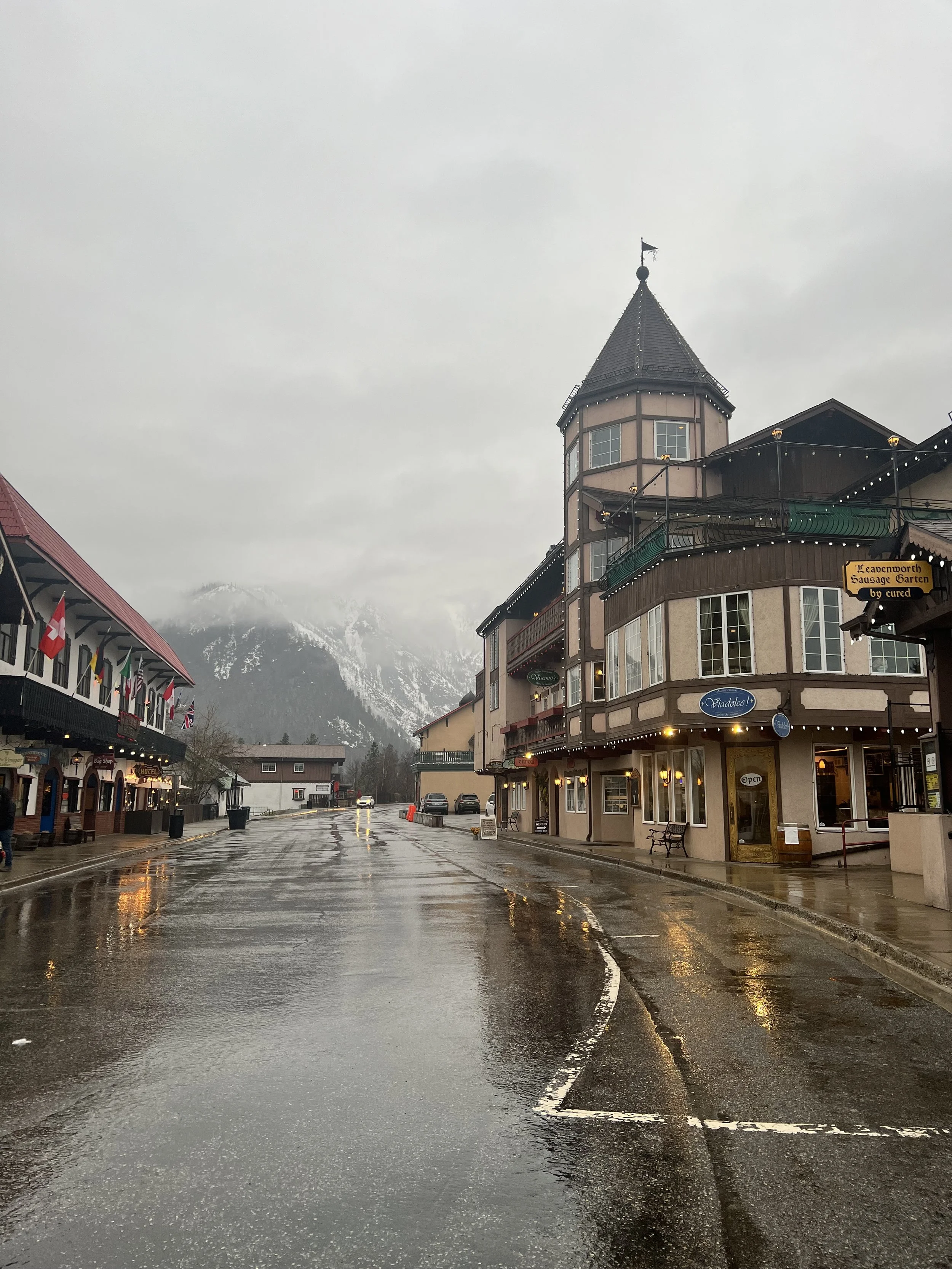 Rainy street scene in a small town with historic buildings and snow-capped mountains in the background.