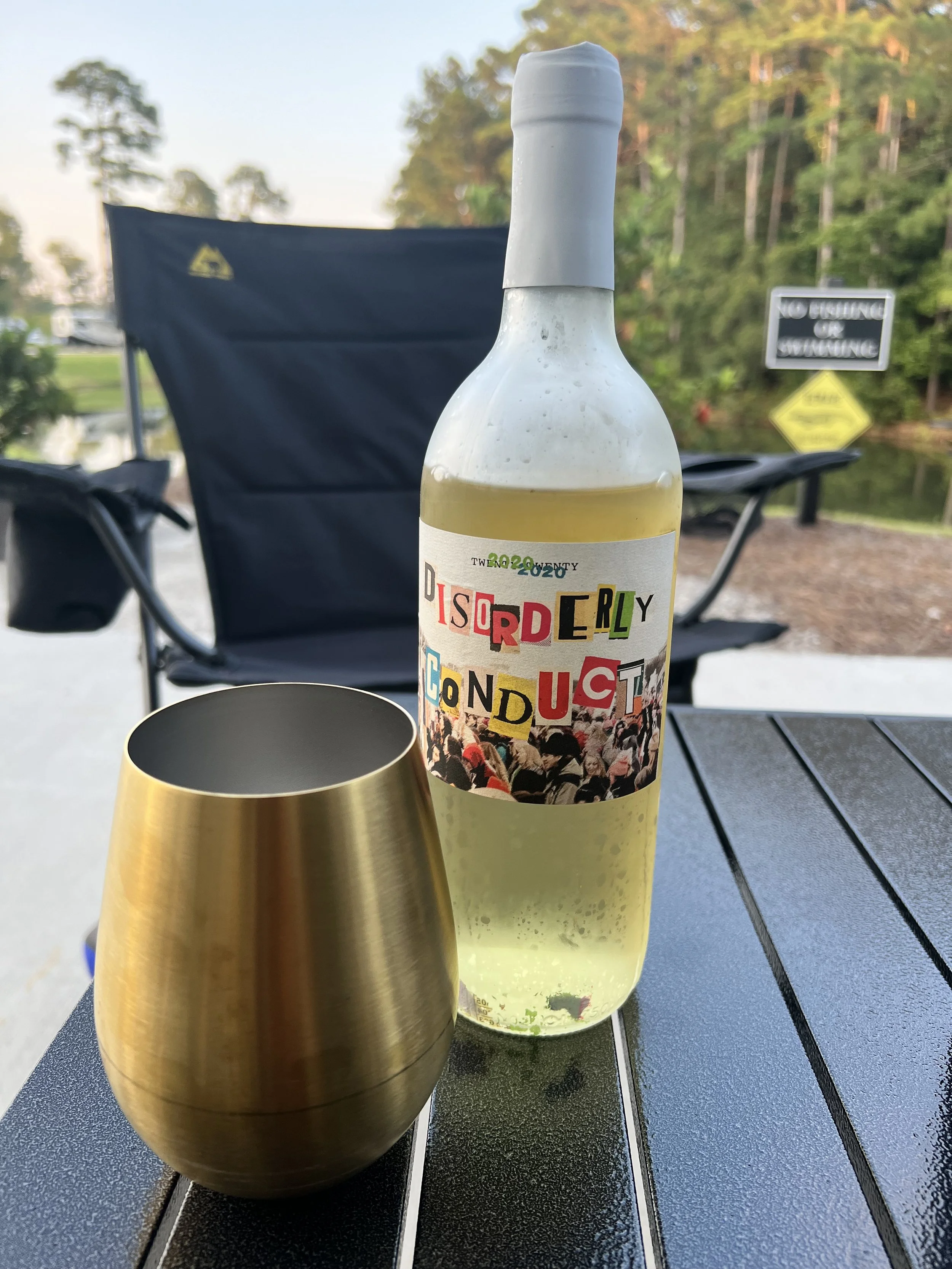 A bottle of white wine with a label reading 'Disorderly Conduct' and a golden metallic wine glass on a black outdoor table, with a background of trees, a lake, and a sign that says 'No fishing or swimming'.