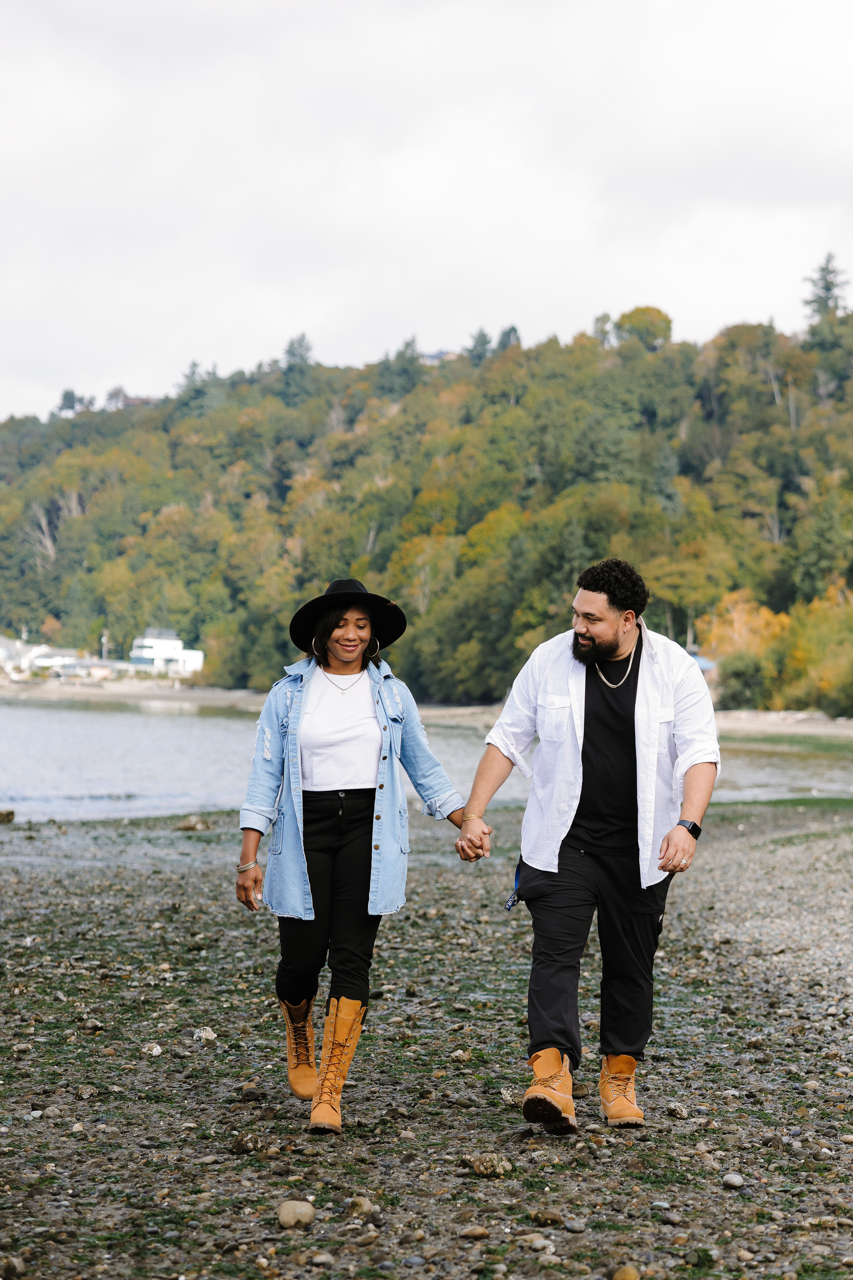 A couple walking hand-in-hand on a rocky beach near the water, surrounded by trees with fall foliage.