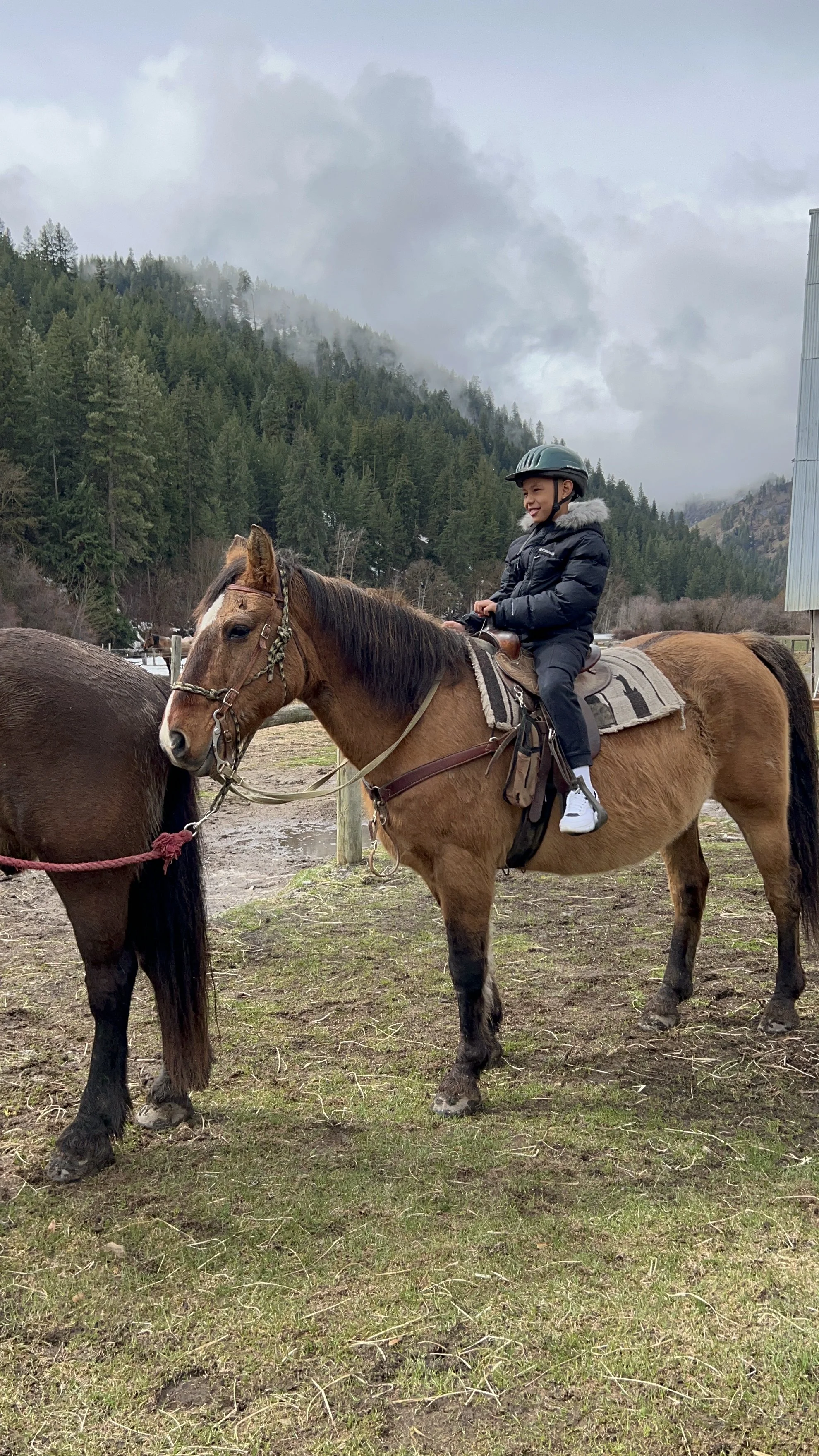 A young boy wearing a black jacket and helmet is riding a brown horse with a saddle, while a woman holds the horse's reins in a rural, mountainous area with green hills and cloudy sky.