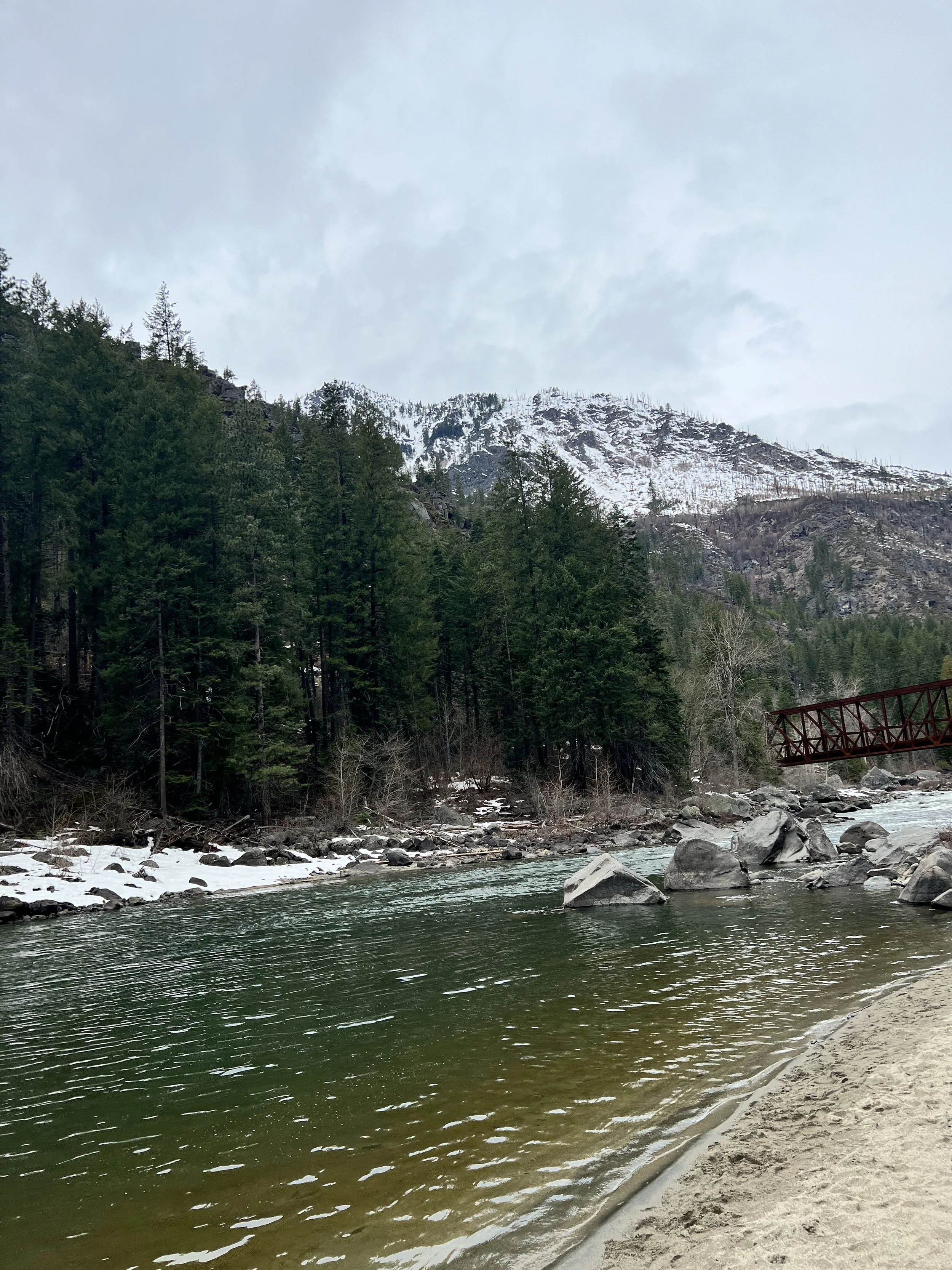 A scenic river flowing through a forested mountain area with snow on the ground and mountain peaks in the background, and a small red bridge over the water.