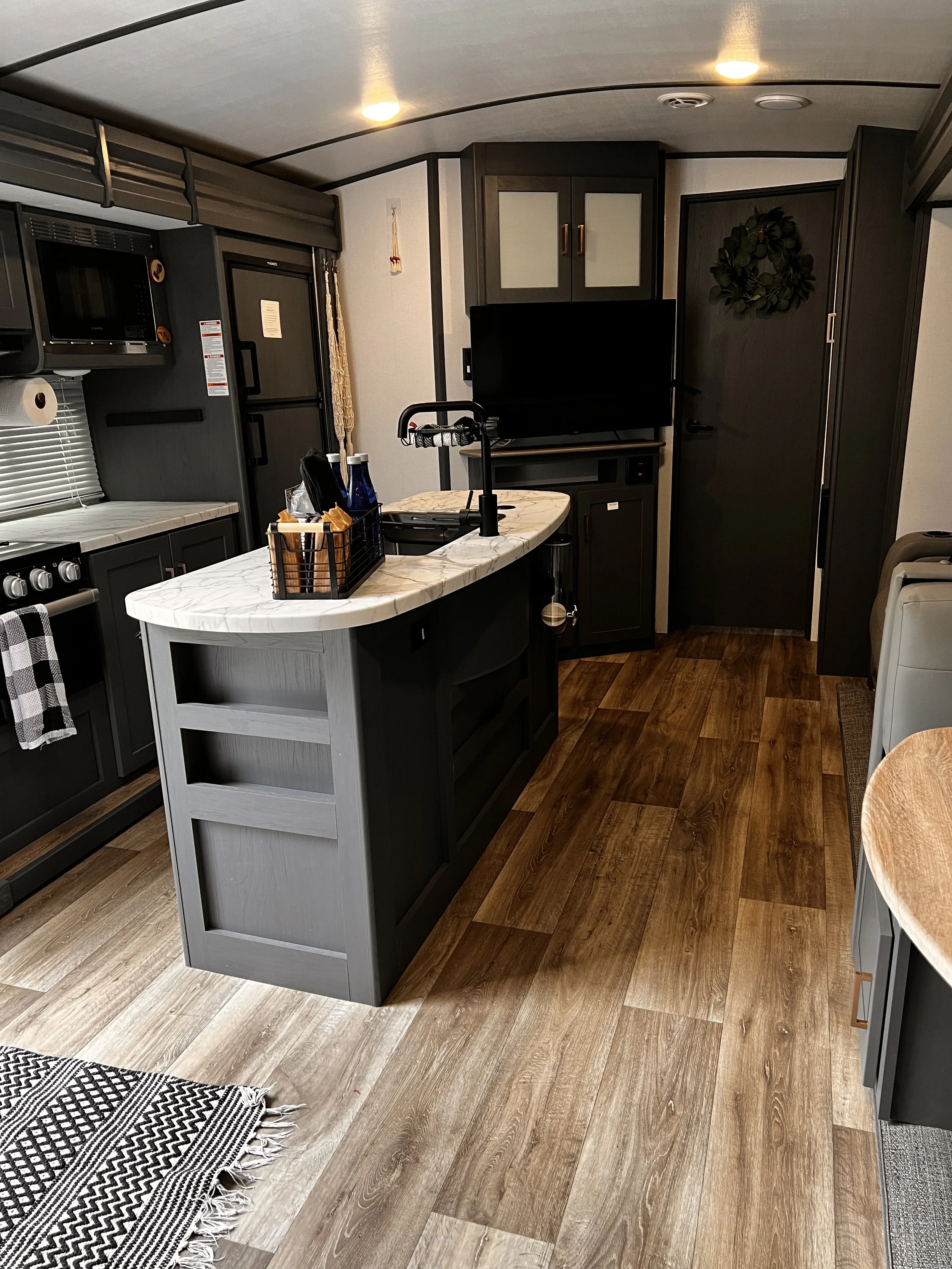 Interior of a modern kitchen in a recreational vehicle with dark cabinetry, a small island with a marble countertop, and wood flooring.