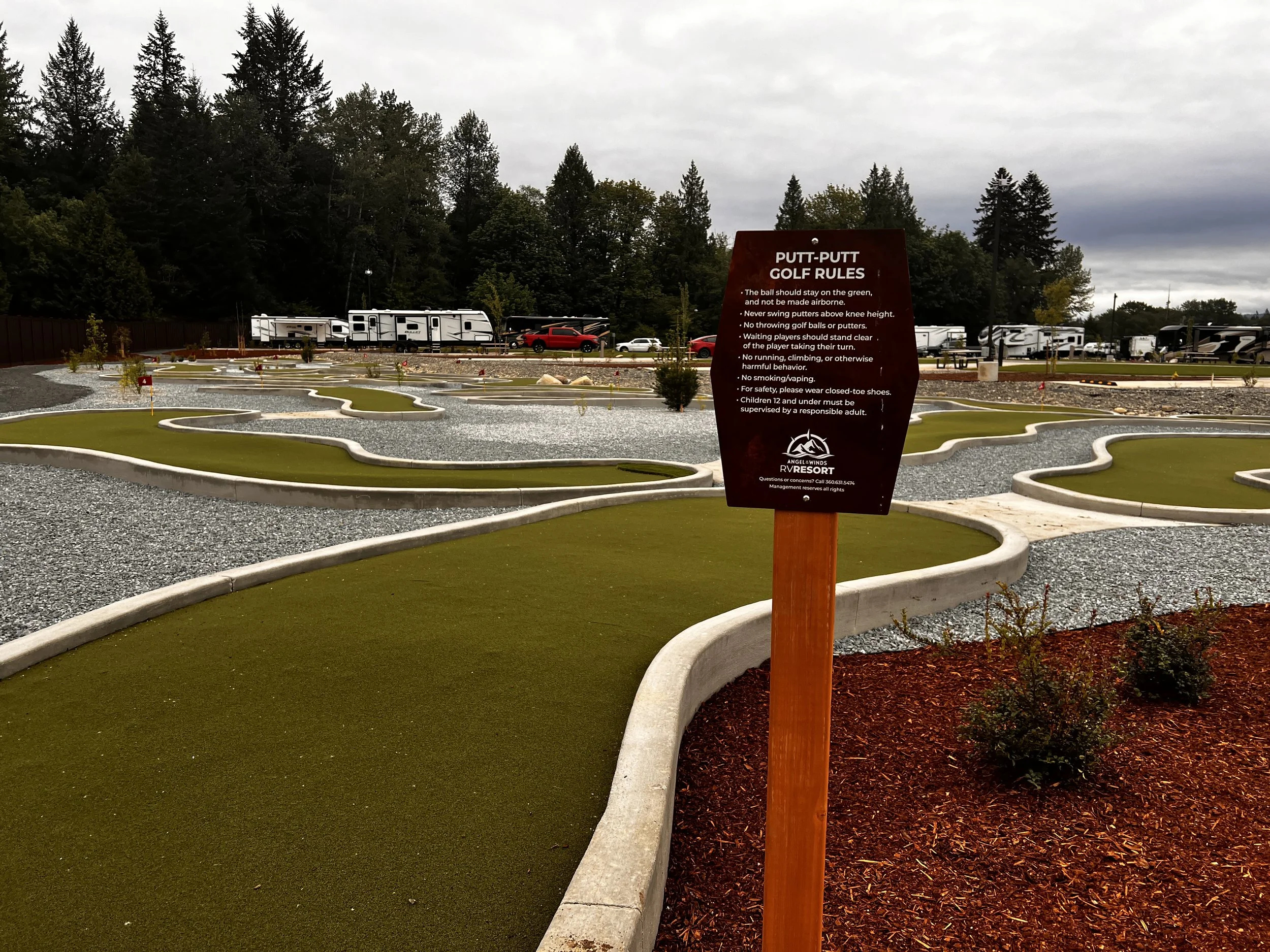 Miniature golf course with winding artificial grass holes, gravel pathways, and a brown sign with rules posted in a campground area with RVs and trees in the background under an overcast sky.