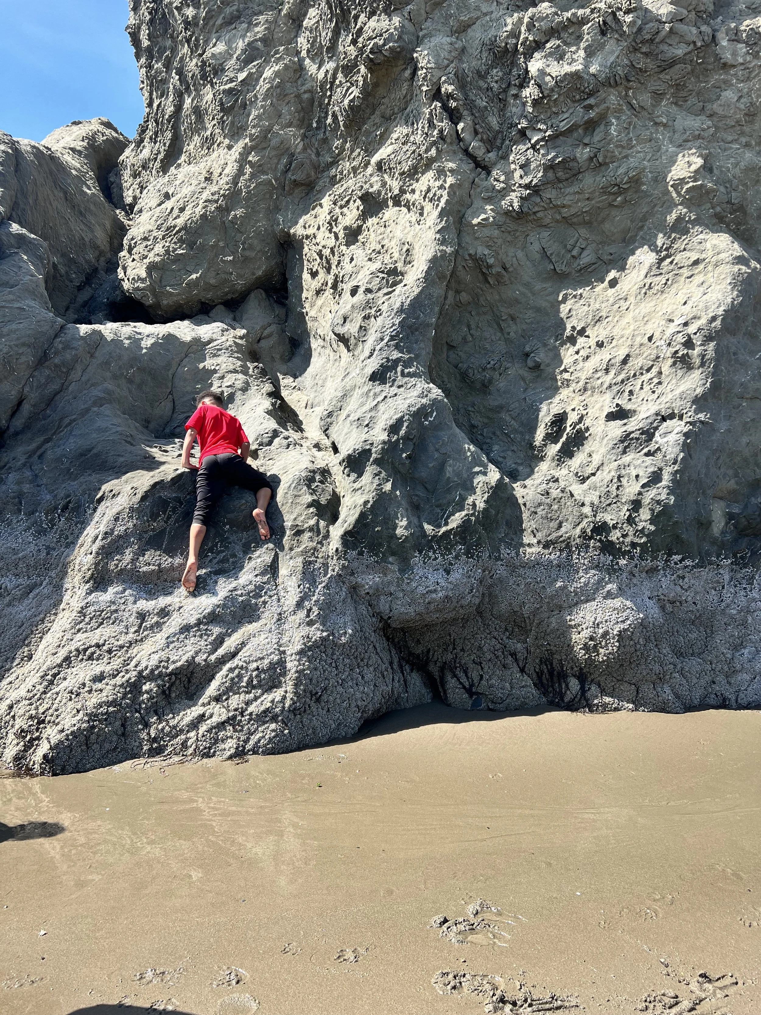 A person climbing a large rocky formation on the beach, with sand in the foreground and clear blue sky above.