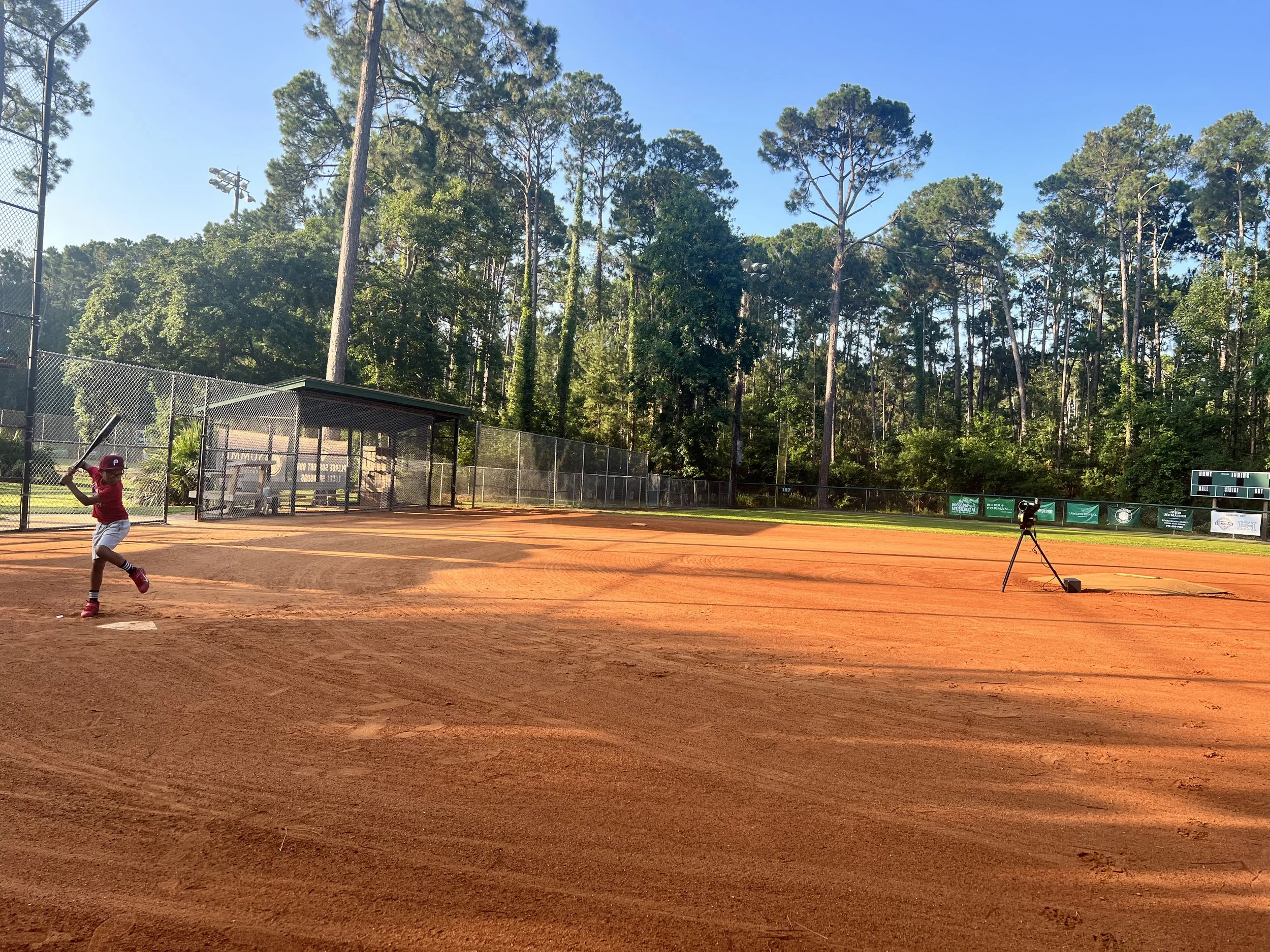 A young baseball player wearing a red uniform and batting helmet is swinging a bat on a clay baseball field. The field is surrounded by a chain-link fence, with a small covered area and a camera on a tripod on the right. Tall trees and a clear blue s