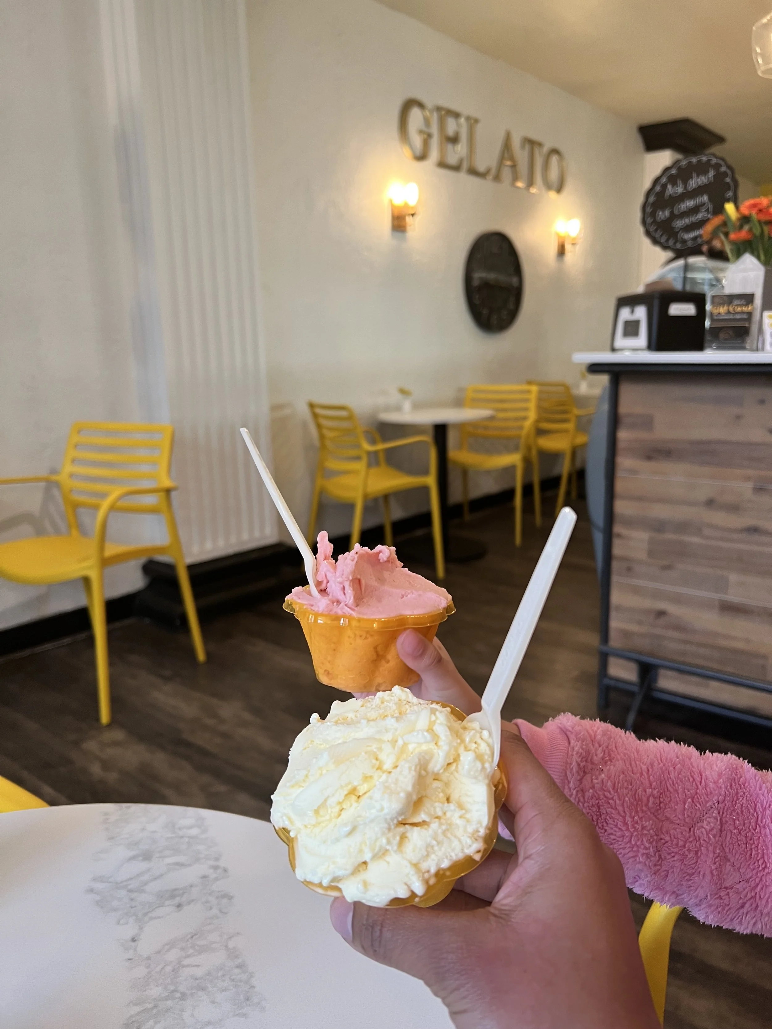 Two cups of ice cream, one with vanilla and the other with strawberry, held by a person in an ice cream shop with yellow chairs and a sign that reads 'GELATO' on the wall.