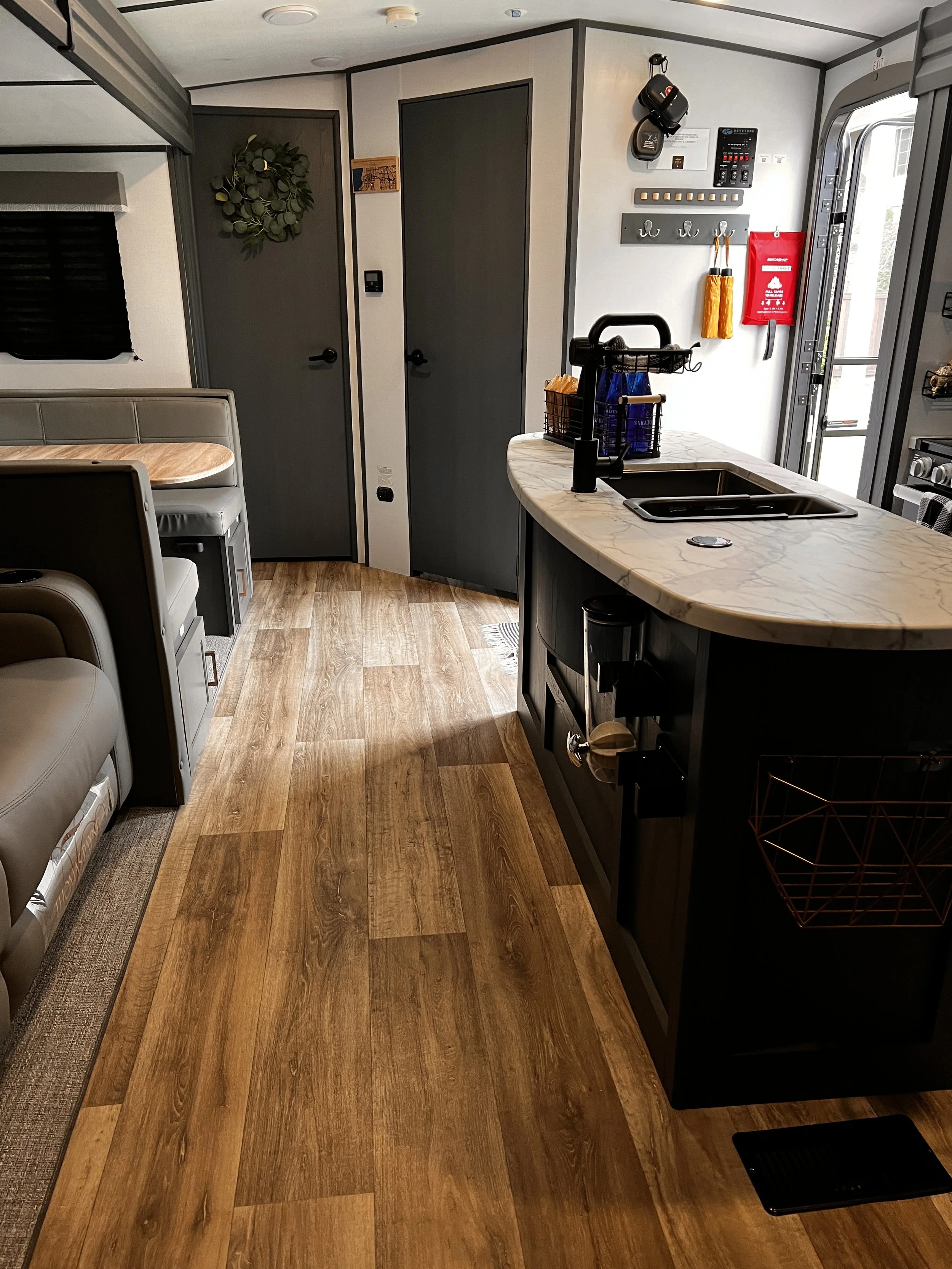Interior of a tiny home with wooden flooring, a kitchen island with a marble countertop, sink, and dishwasher, a door with a wreath, and various wall hooks and control panels near the entry door.
