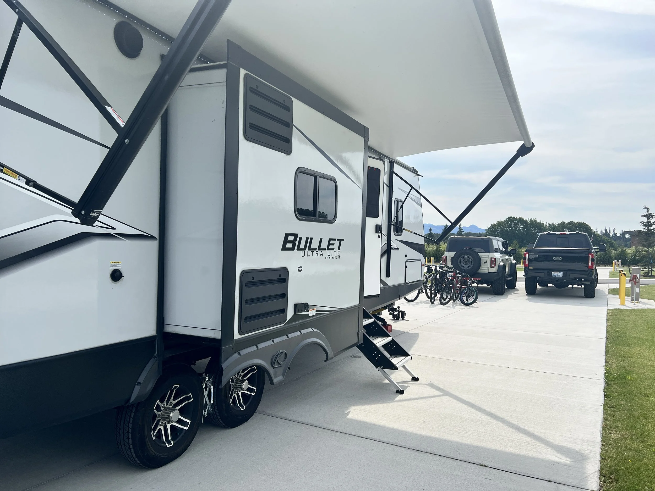 A white camping trailer with the words 'Bullet Ultra Lite' on its side, parked under a large extended canopy, with two black pickup trucks and several bicycles in the background.